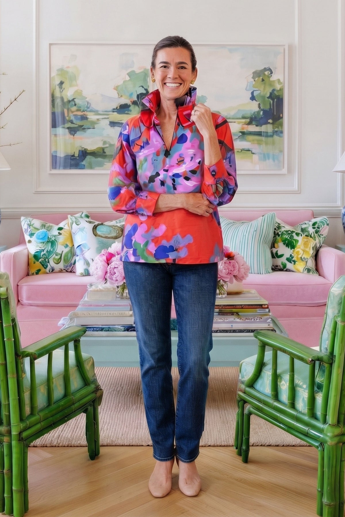 A woman stands in a bright living room wearing a colorful floral long-sleeve button-up shirt with blue jeans. The room features a pink sofa with patterned pillows, green bamboo-style chairs, and a framed landscape painting on the wall.