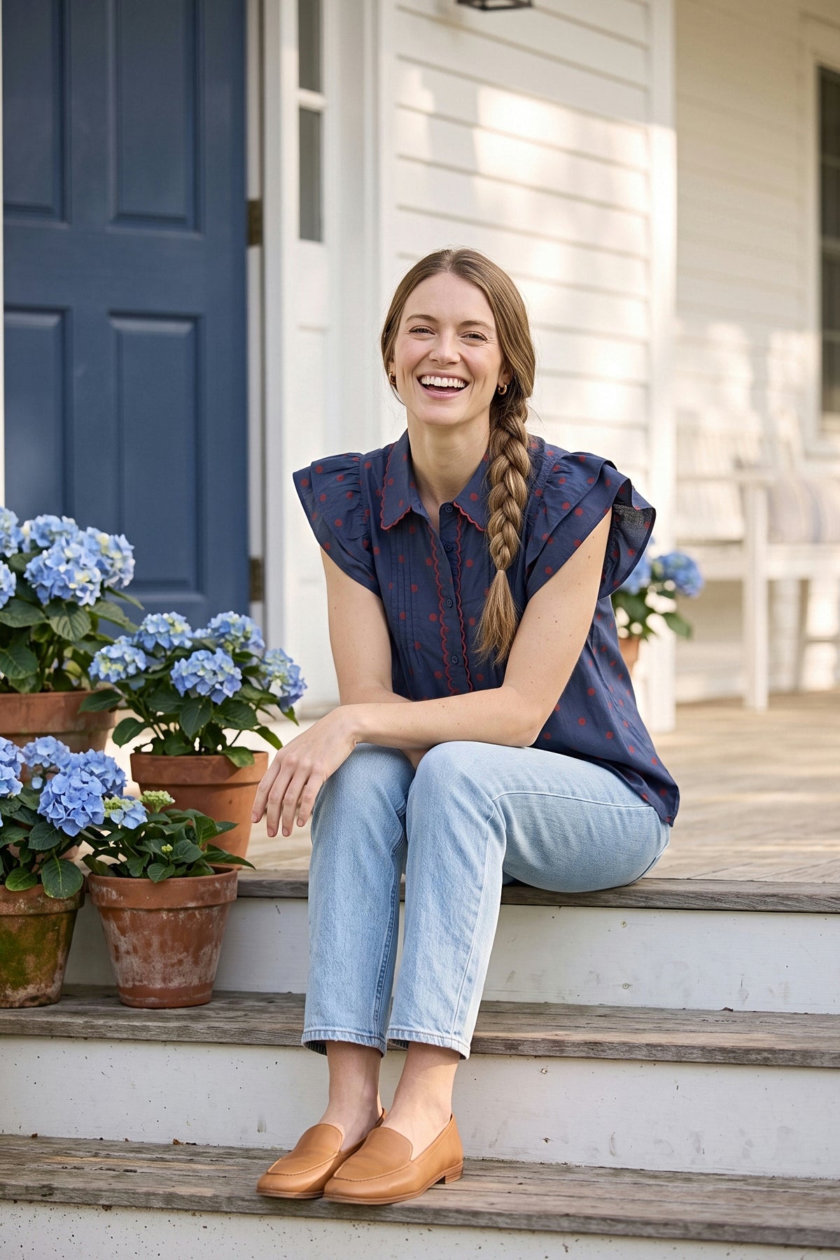 A woman laughs on porch steps in the navy polka dot blouse with red scalloped trim and flutter sleeves.