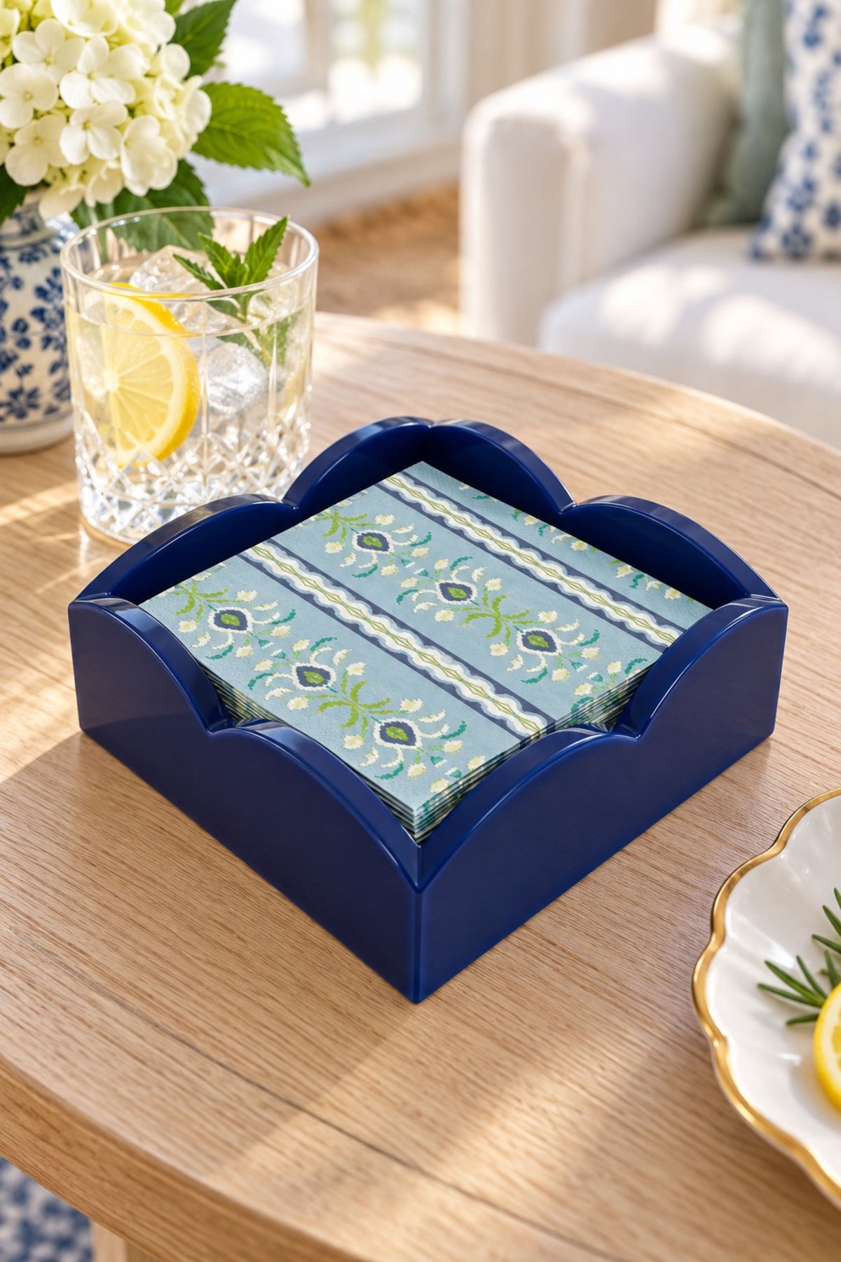 A navy blue ceramic coasters holder filled with patterned napkins, styled on a light wood table next to a glass of lemonade in a bright, sunlit room.