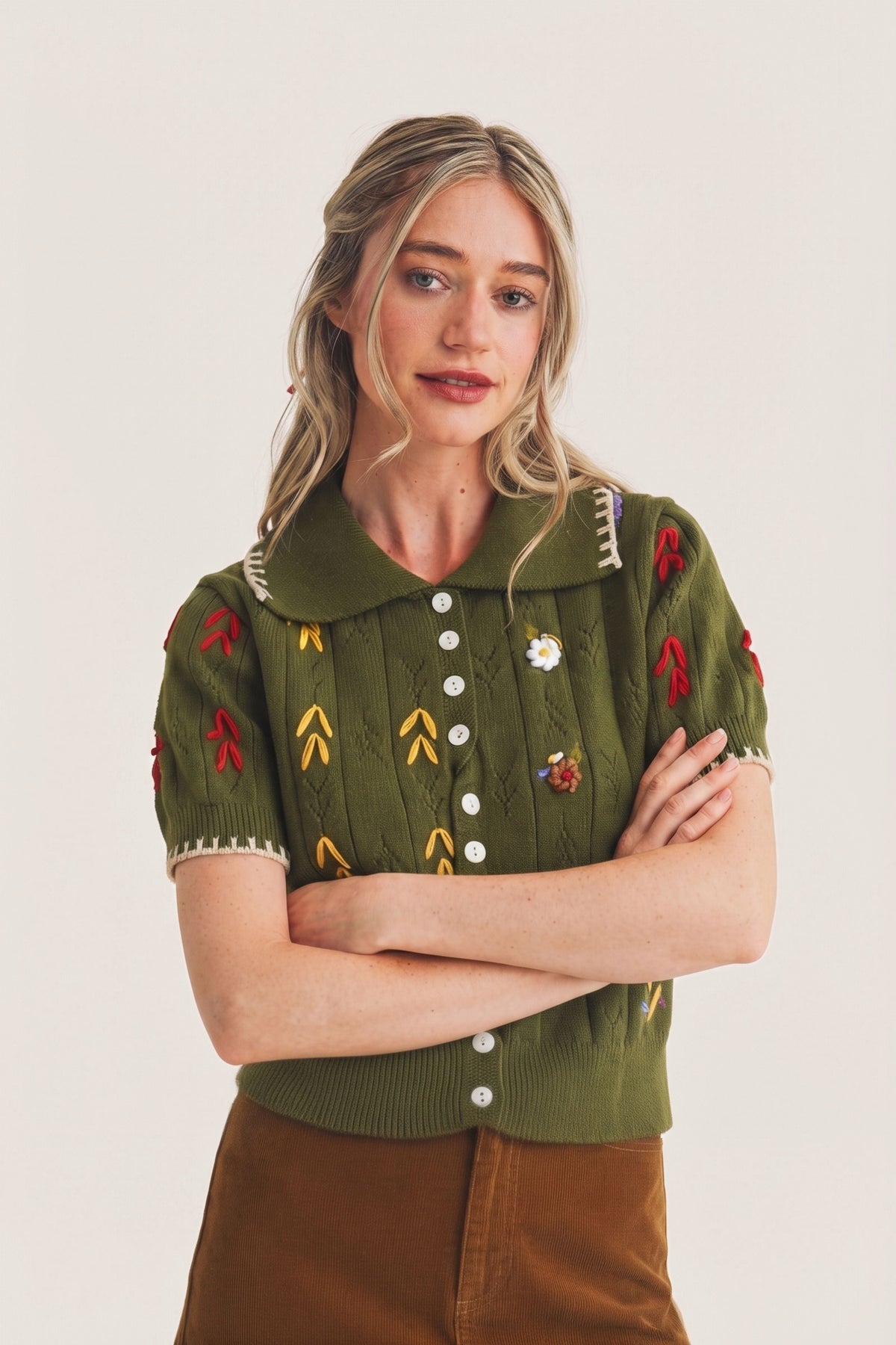 A woman modeling a short-sleeve olive green knit button-front top with embroidered red and yellow motifs, a pointed collar, and white buttons, paired with brown pants against a plain light background.