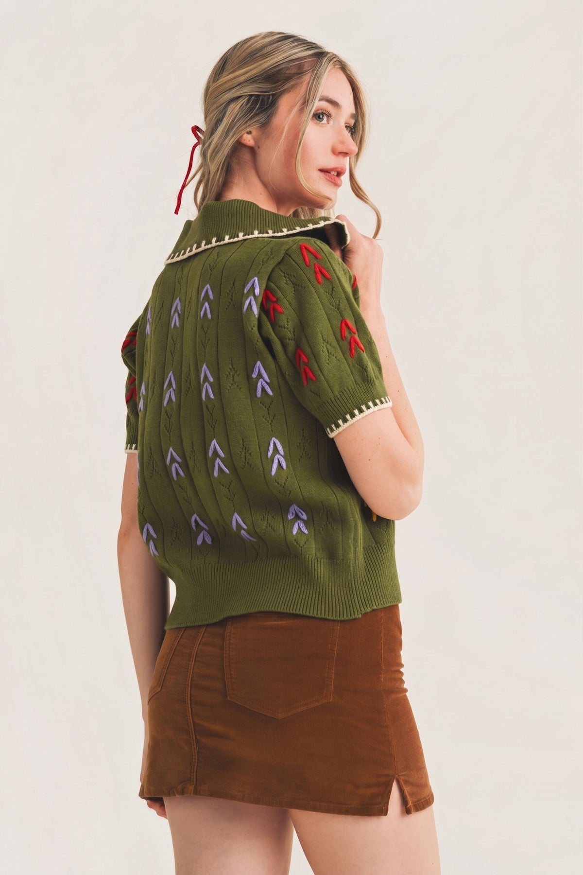 A woman wearing a short-sleeve olive green knit sweater with red and purple embroidered arrow motifs and a brown mini skirt, photographed against a plain light background.