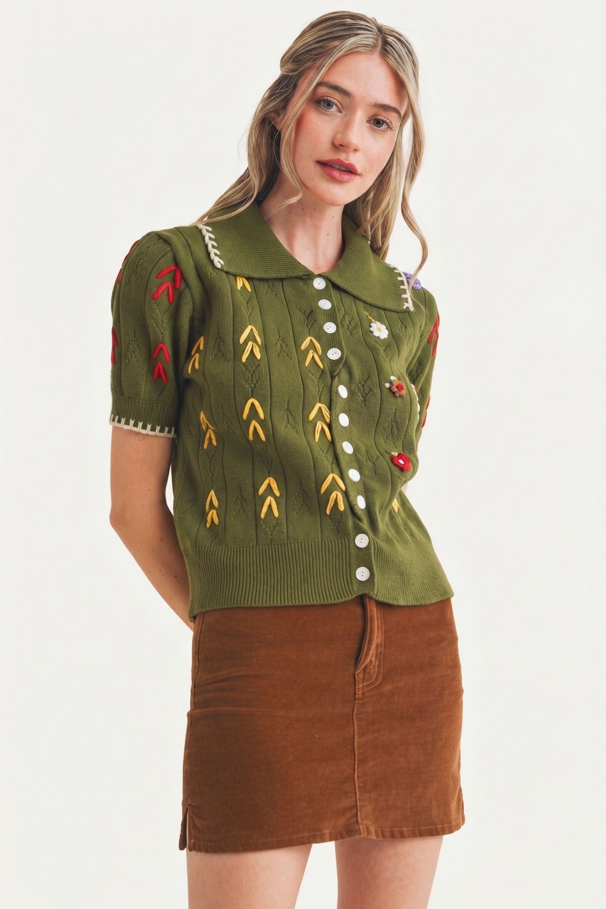 A woman modeling a short-sleeve olive green knit top with colorful embroidered motifs and a brown corduroy mini skirt, photographed against a plain light background.