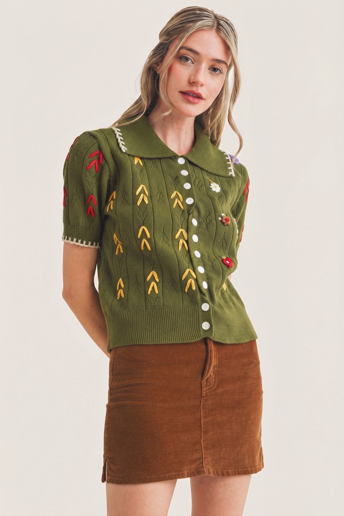 A woman modeling a short-sleeve olive green knit top with colorful embroidered motifs and a brown corduroy mini skirt, photographed against a plain light background.