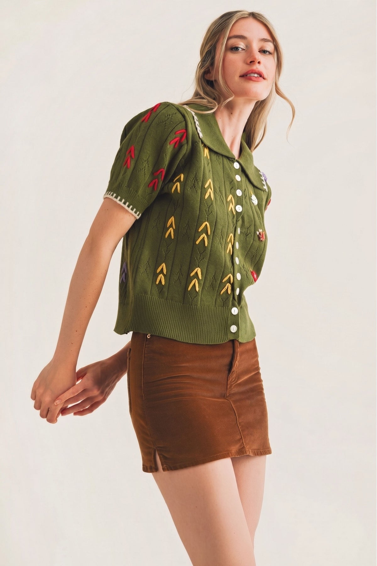 A woman modeling a short-sleeve olive green knit button-up top with colorful embroidered arrow motifs and a brown mini skirt, set against a plain light beige background.