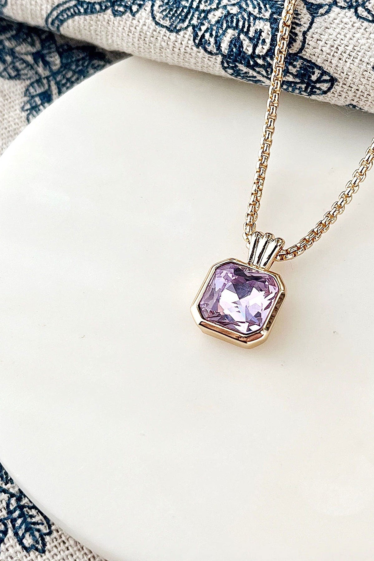 The lavender cushion-cut crystal pendant and gold box chain of the Orchid Necklace on white marble.