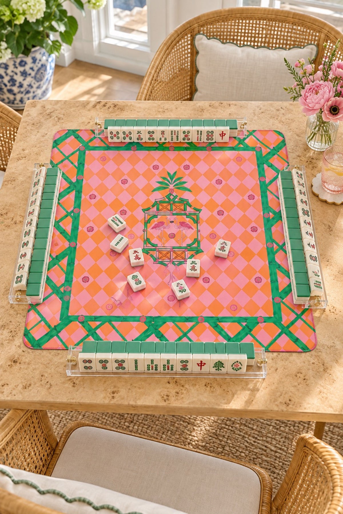 A colorful Mahjong-style tile game set arranged on a light wood dining table in a bright room with wicker chairs and flowers. The mat has a pink and orange diamond pattern with green borders, and several tiles are placed face up in the center.