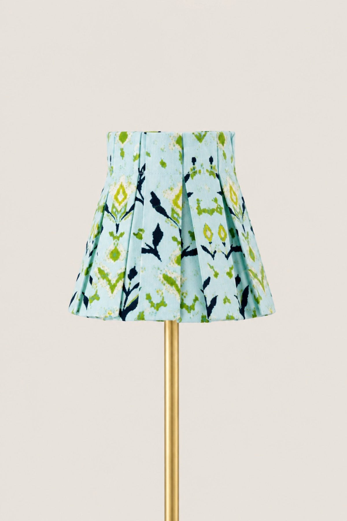 A pleated fabric lampshade with a light blue background and a green, yellow, and black botanical pattern, mounted on a gold lamp stand against a solid light beige background.