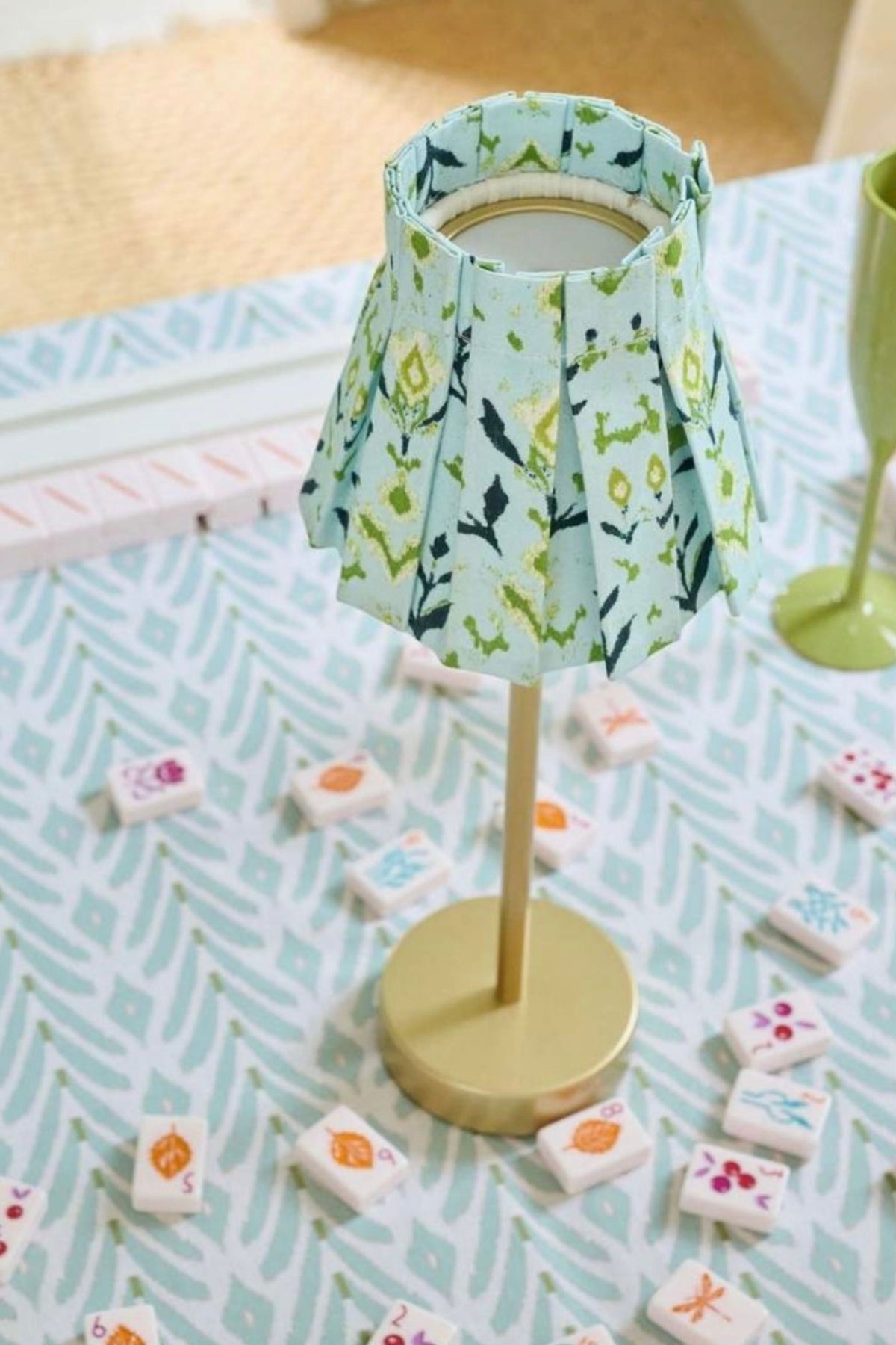 A small decorative table lamp with a pleated light blue and green floral fabric shade and a round gold base, styled on a patterned surface with small white tiles scattered around it.