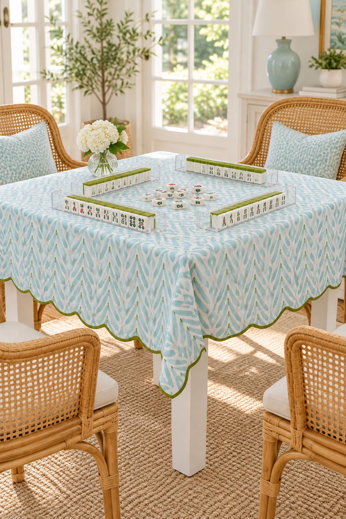 A light blue and white patterned tablecloth with a green scalloped border is draped over a dining table in a bright, sunlit room with wicker chairs and a game of mahjong on top.