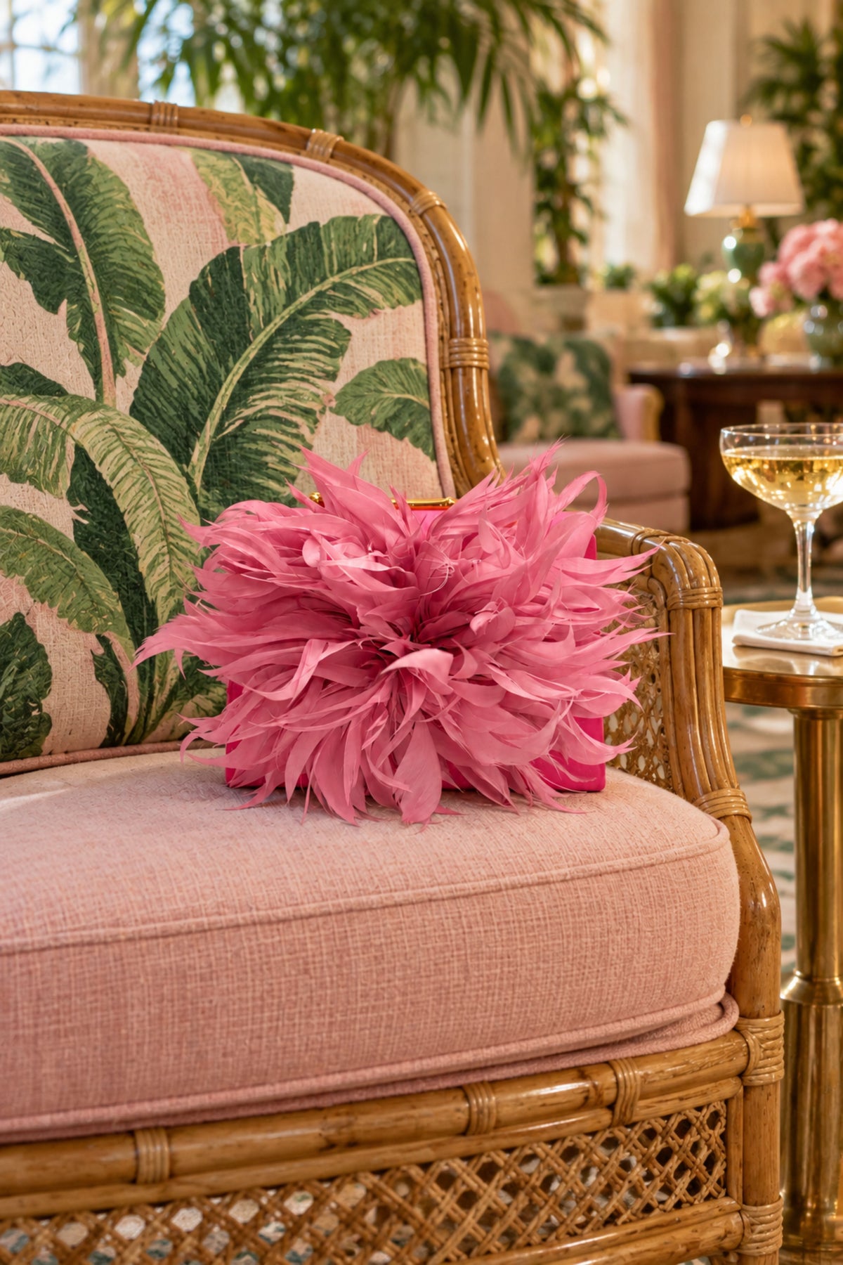 A pink ruffled decorative pillow rests on the cushion of a pink upholstered rattan armchair with a green tropical leaf patterned backrest, set in a softly lit indoor room.
