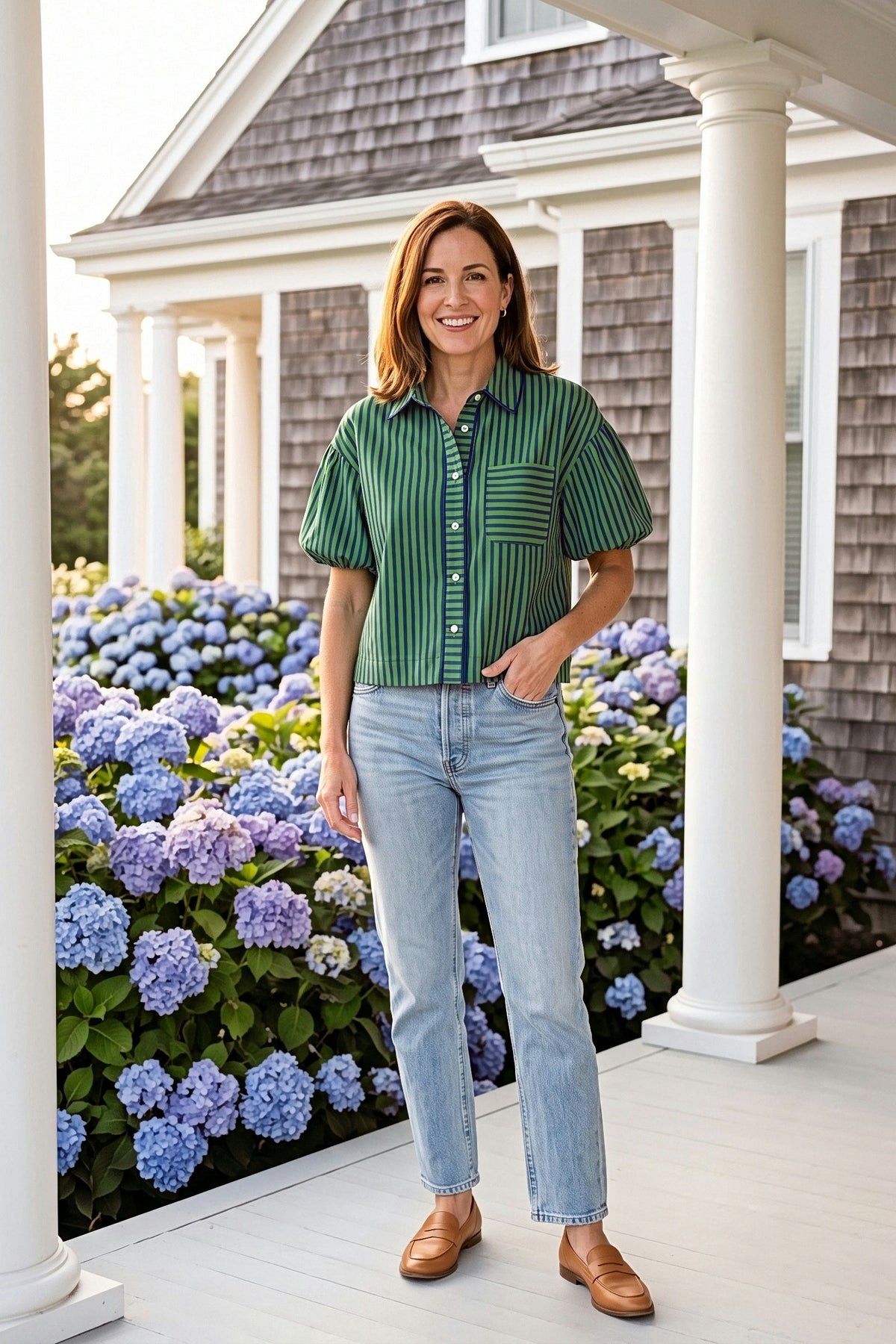 A woman on a white porch with blue hydrangeas wearing the Preppy navy and green stripe top with light jeans and tan loafers.