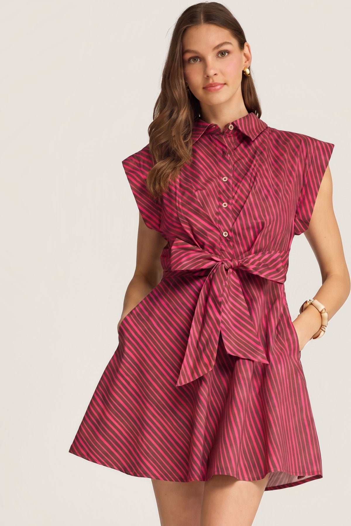 A woman models a cap sleeve collared shirt dress with a tie waist and a raspberry and brown diagonal stripe pattern, set against a plain light background.