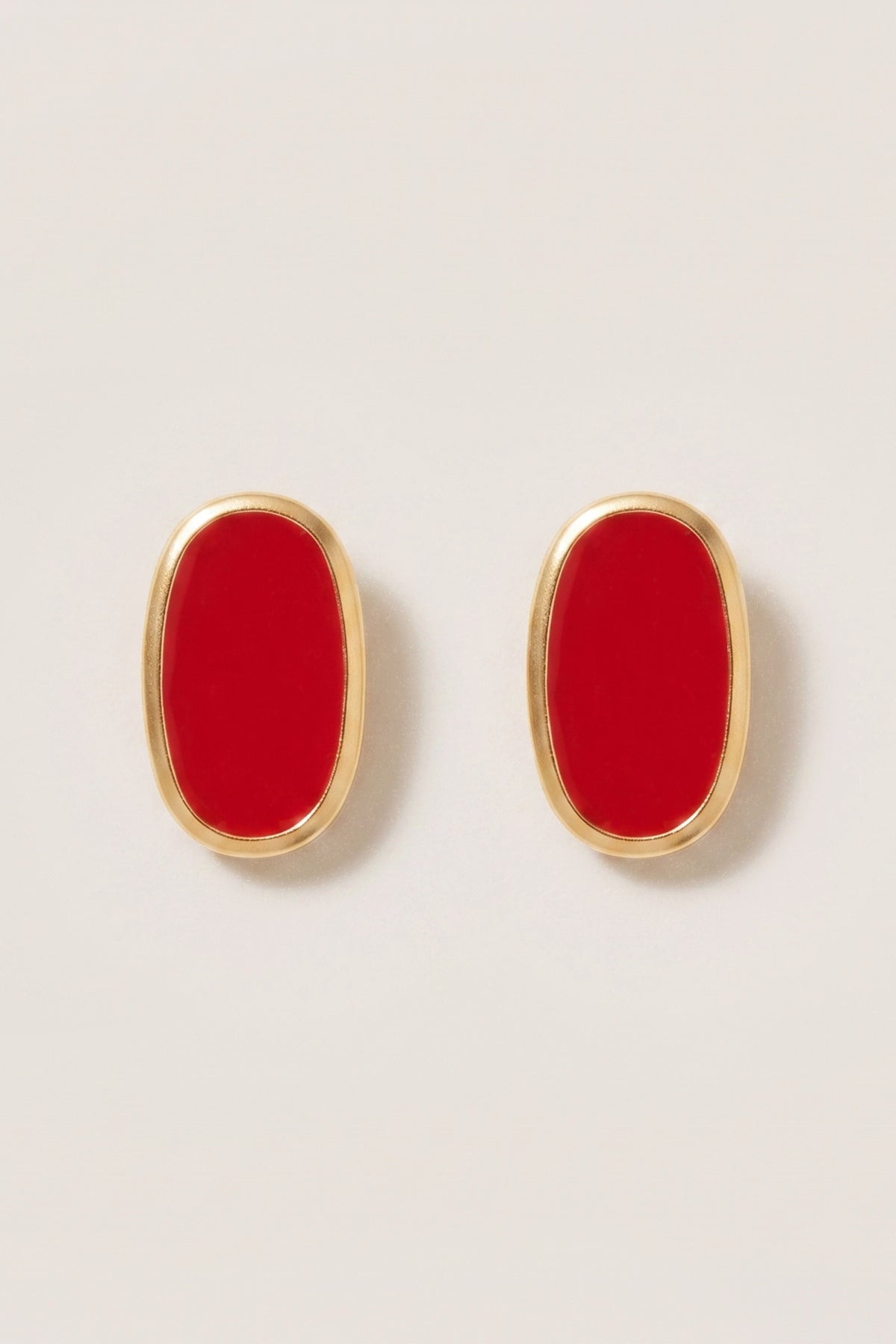 A pair of gold oval stud earrings with vibrant red enamel centers, displayed on a clean, light beige background.