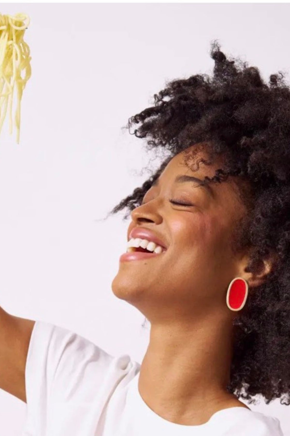 A smiling woman with curly dark hair looks upward while wearing a white top and a bright red oval earring. A small portion of yellow pasta is visible in the upper left against a plain light background.