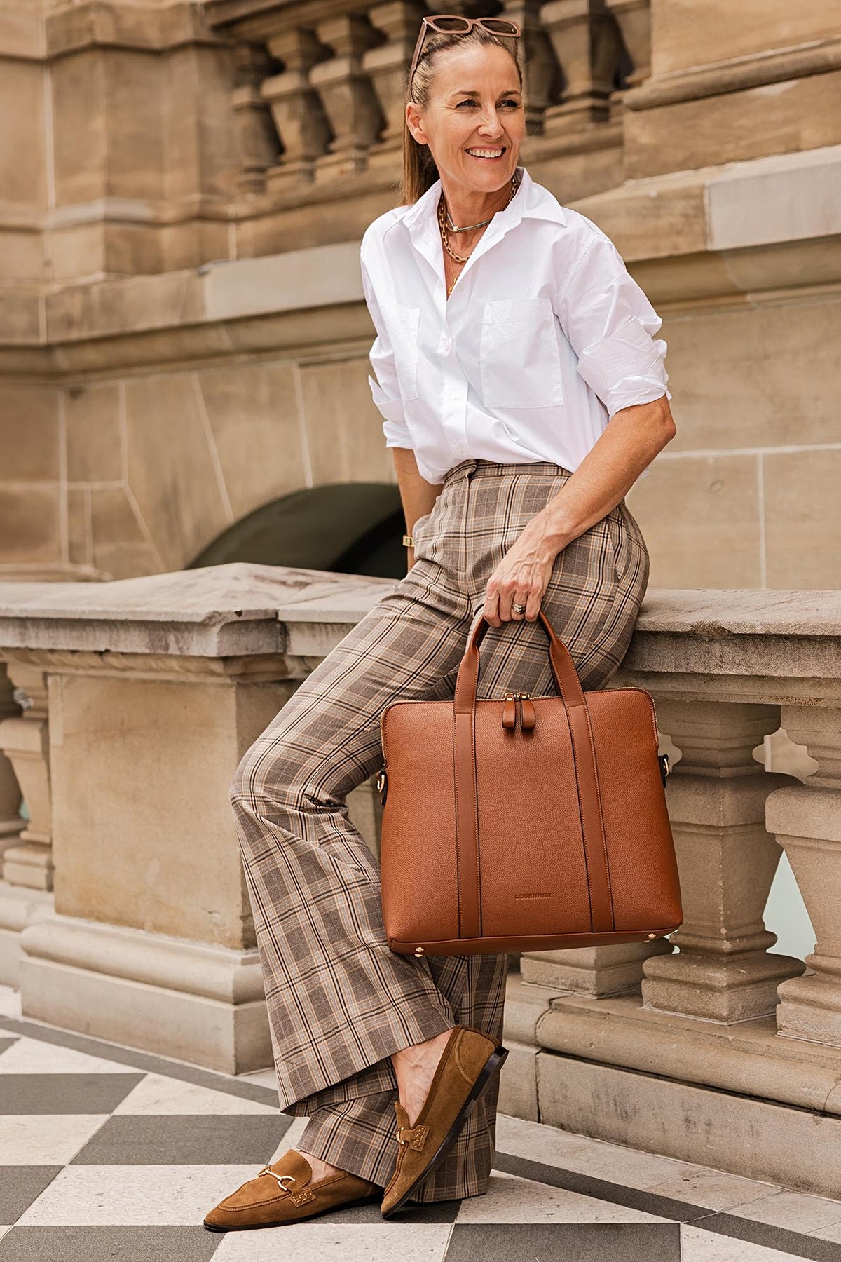 A woman in a white shirt and plaid trousers seated with the tan Rhodes Laptop Bag on her knee.