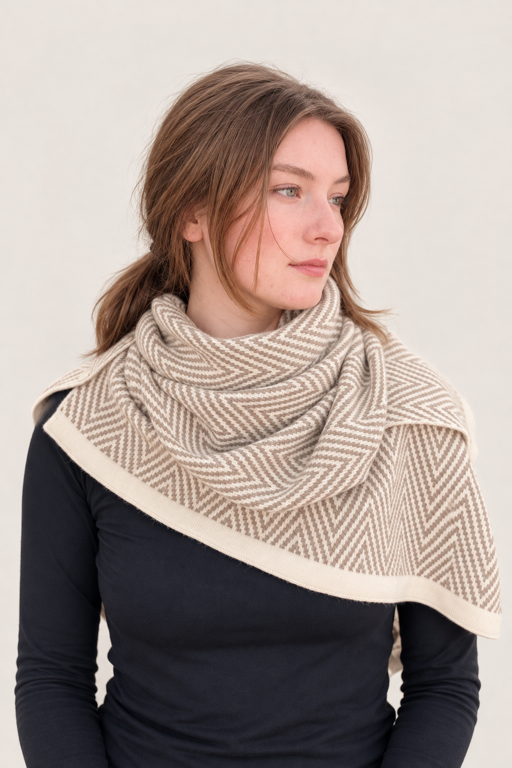A woman wearing a beige and cream herringbone patterned wrap or scarf draped over her shoulders, paired with a black long-sleeve top, against a solid light beige background.
