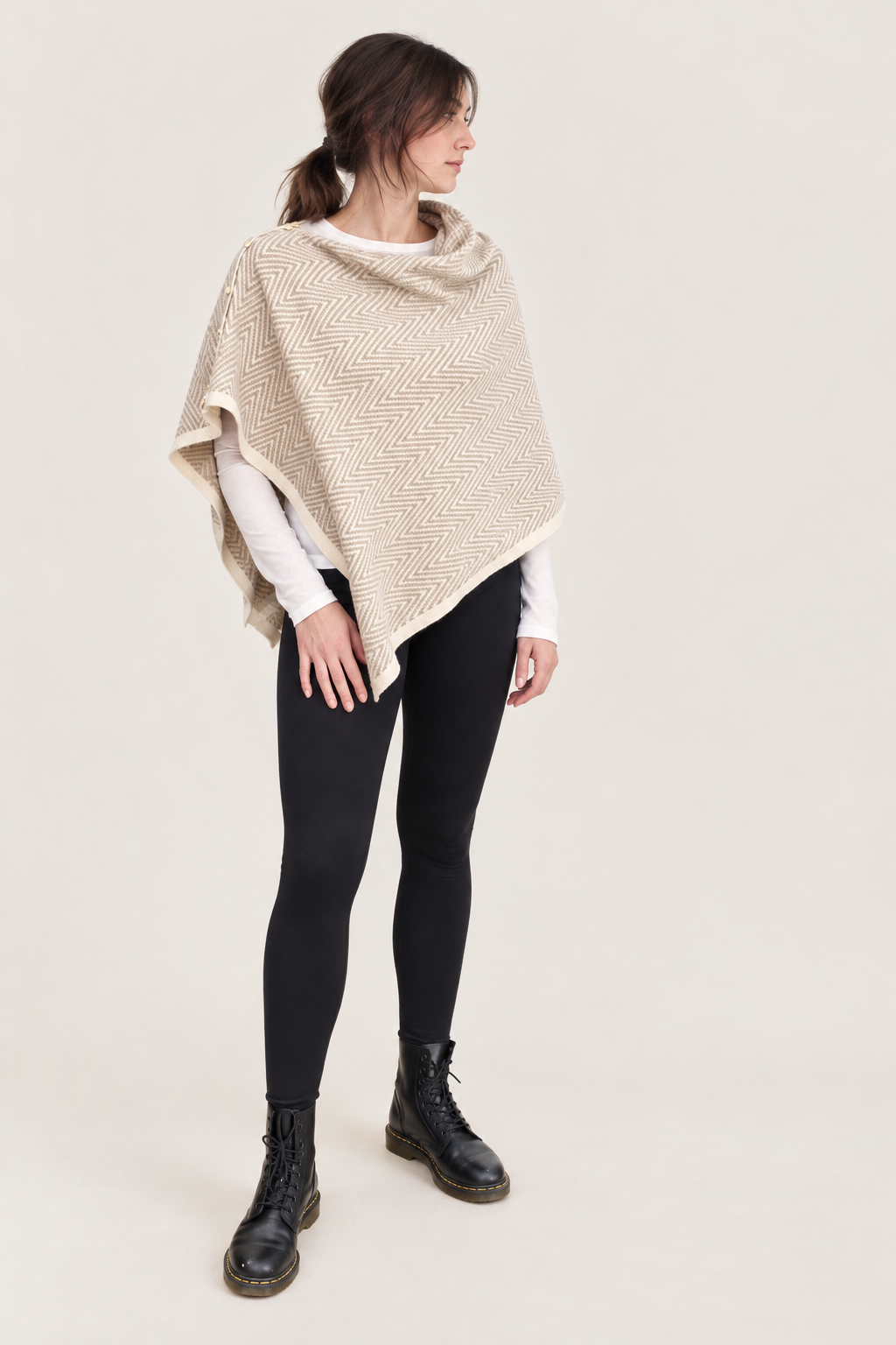 A woman modeling a chunky knit beige poncho over a white long-sleeve top, black leggings, and black lace-up boots, set against a solid light beige background.