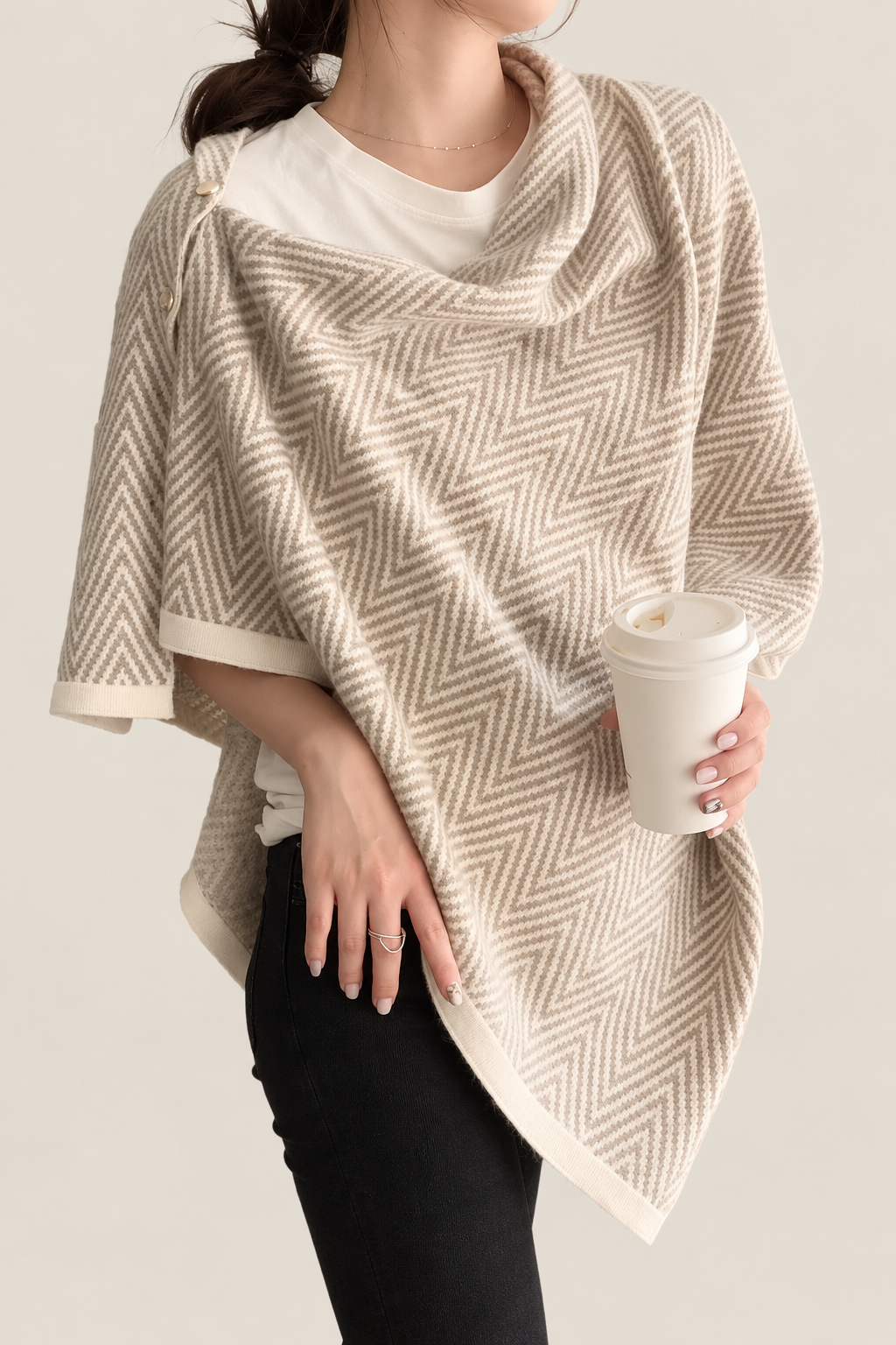 A woman wearing a beige and cream herringbone-patterned knit wrap poncho over a white t-shirt and black pants, holding a white takeaway coffee cup against a light beige background.
