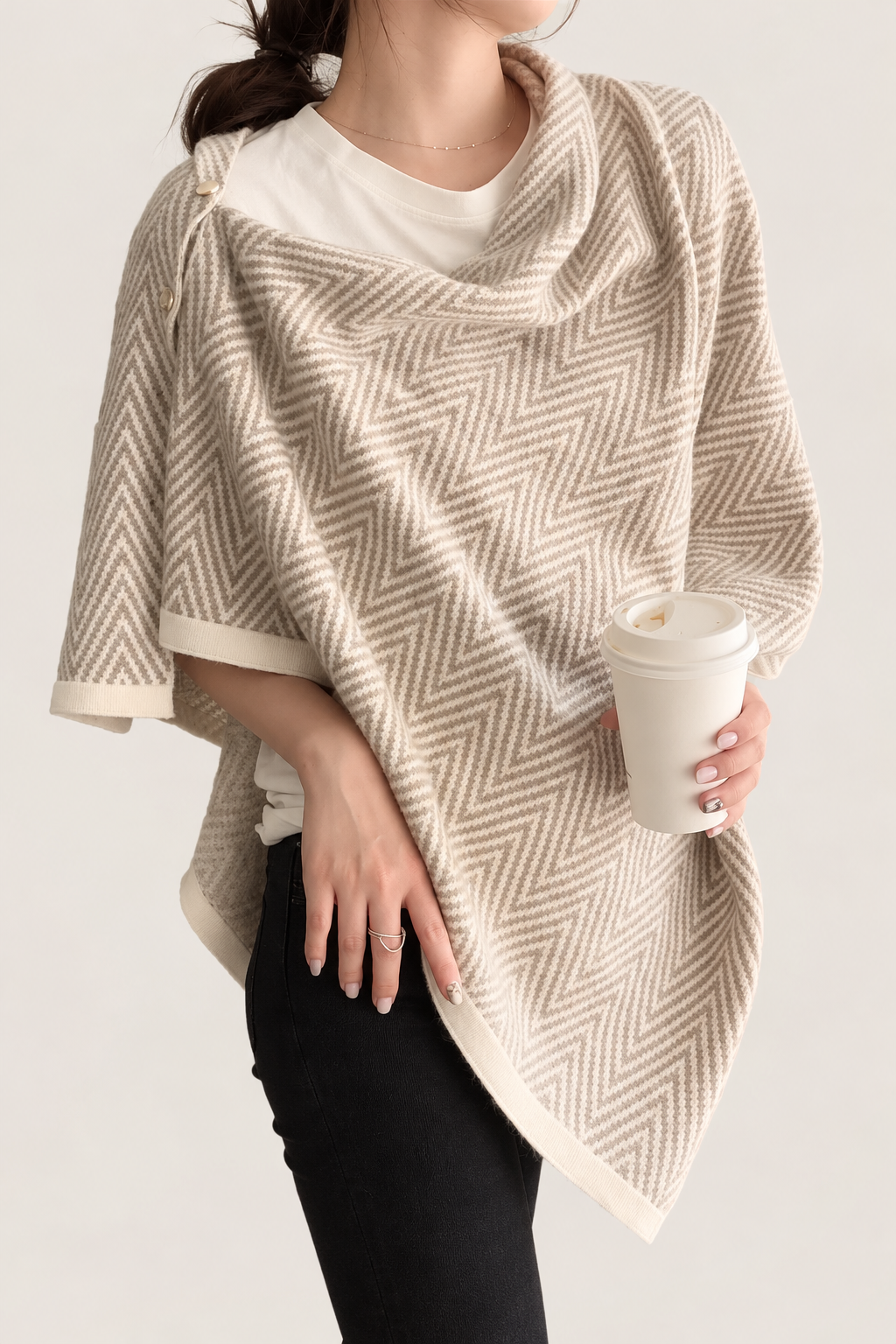 A woman wearing a beige and cream herringbone-patterned knit wrap poncho over a white t-shirt and black pants, holding a white takeaway coffee cup against a light beige background.