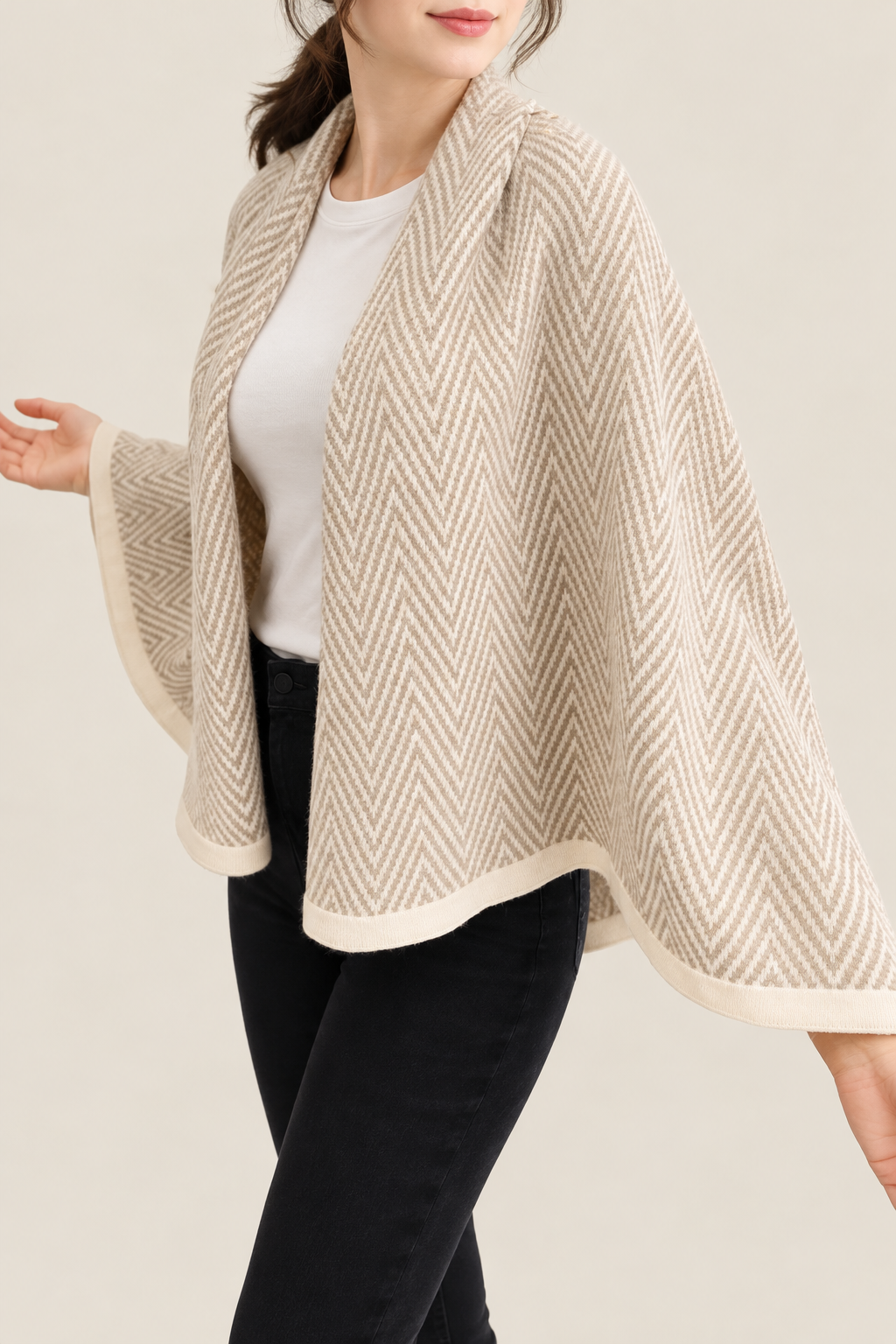 A woman wearing a beige and cream herringbone-patterned knit poncho over a white top and black pants, shown against a plain light beige studio background.