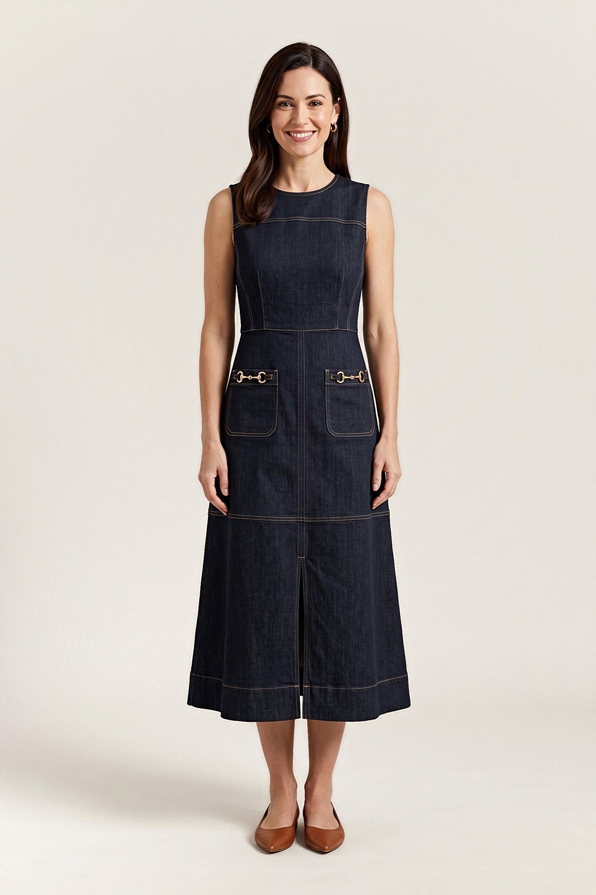 Front view of the sleeveless dark rinse denim midi dress with gold topstitching, horsebit pockets and a front slit.