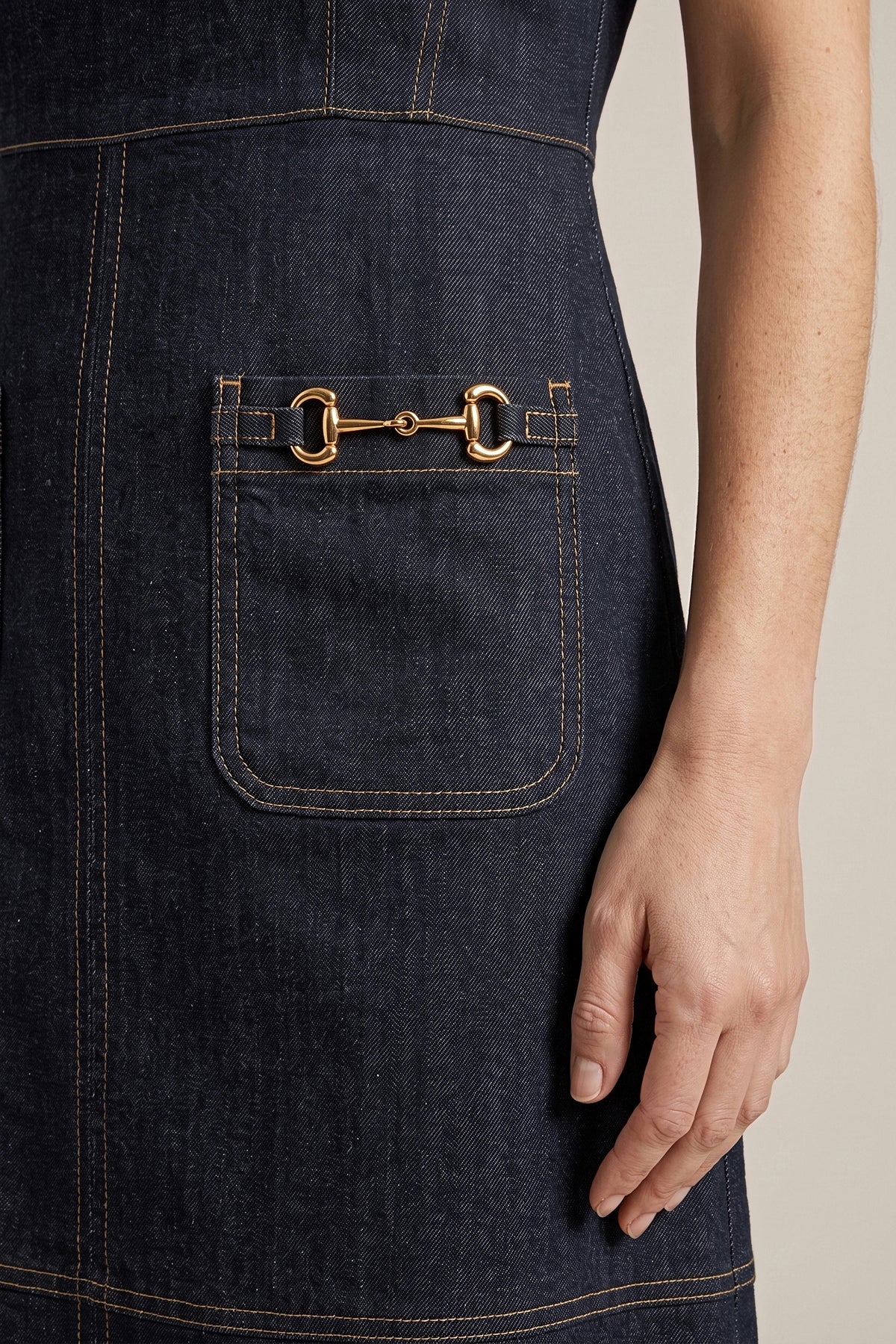 Close-up of the gold horsebit hardware and topstitching on the patch pocket of the dark denim dress.