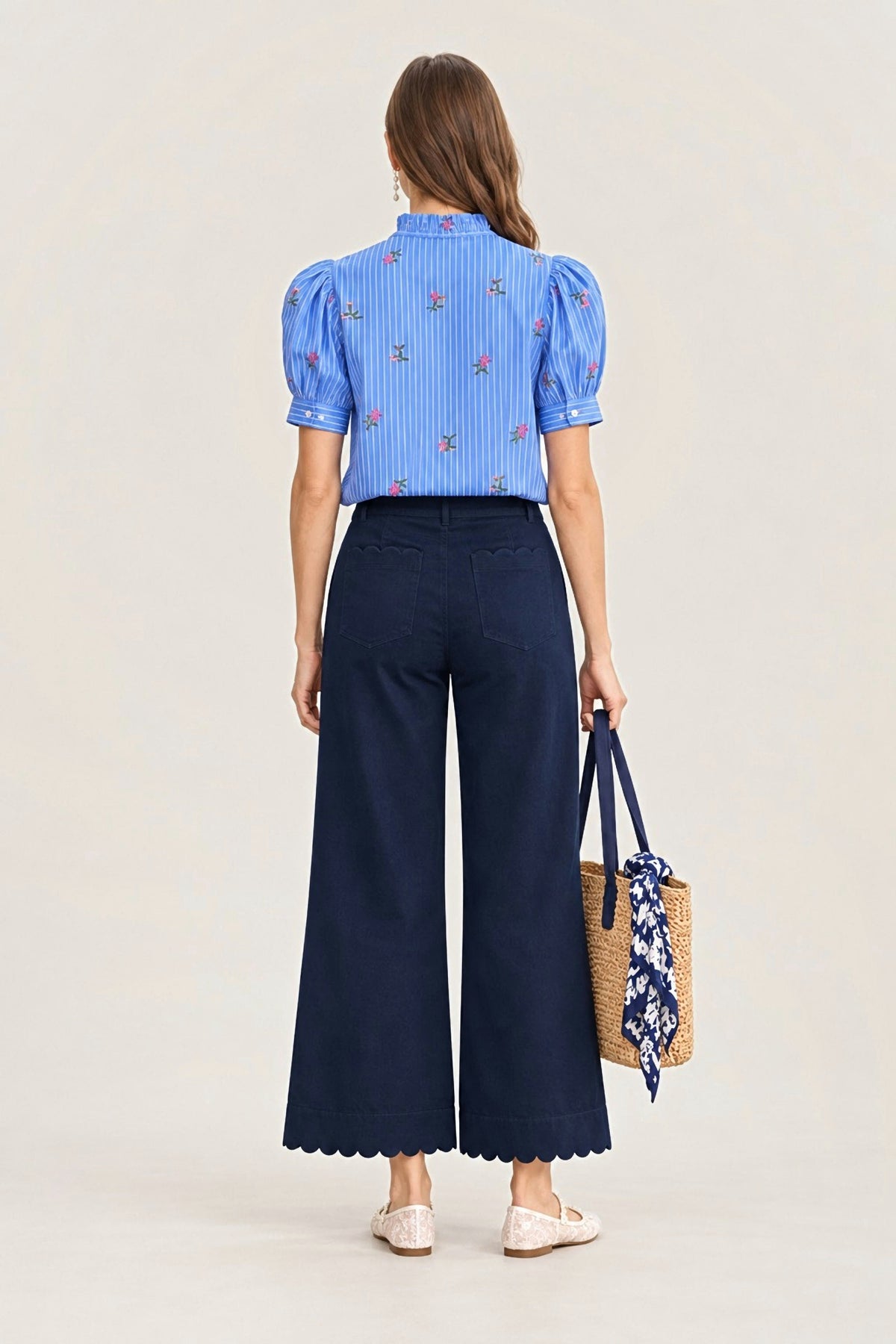 A woman is shown from the back wearing a light blue puff-sleeve blouse with small floral embroidery and dark navy wide-leg pants. She is holding a woven straw tote bag with a blue floral scarf, set against a plain off-white studio background.