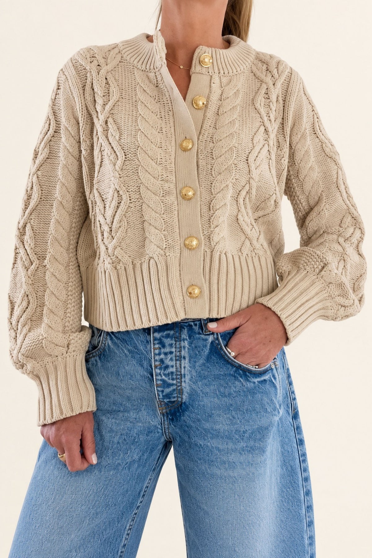 A woman wearing a oatmeal cable-knit cropped cardigan with gold buttons, paired with light-wash blue jeans, against a plain light beige background.