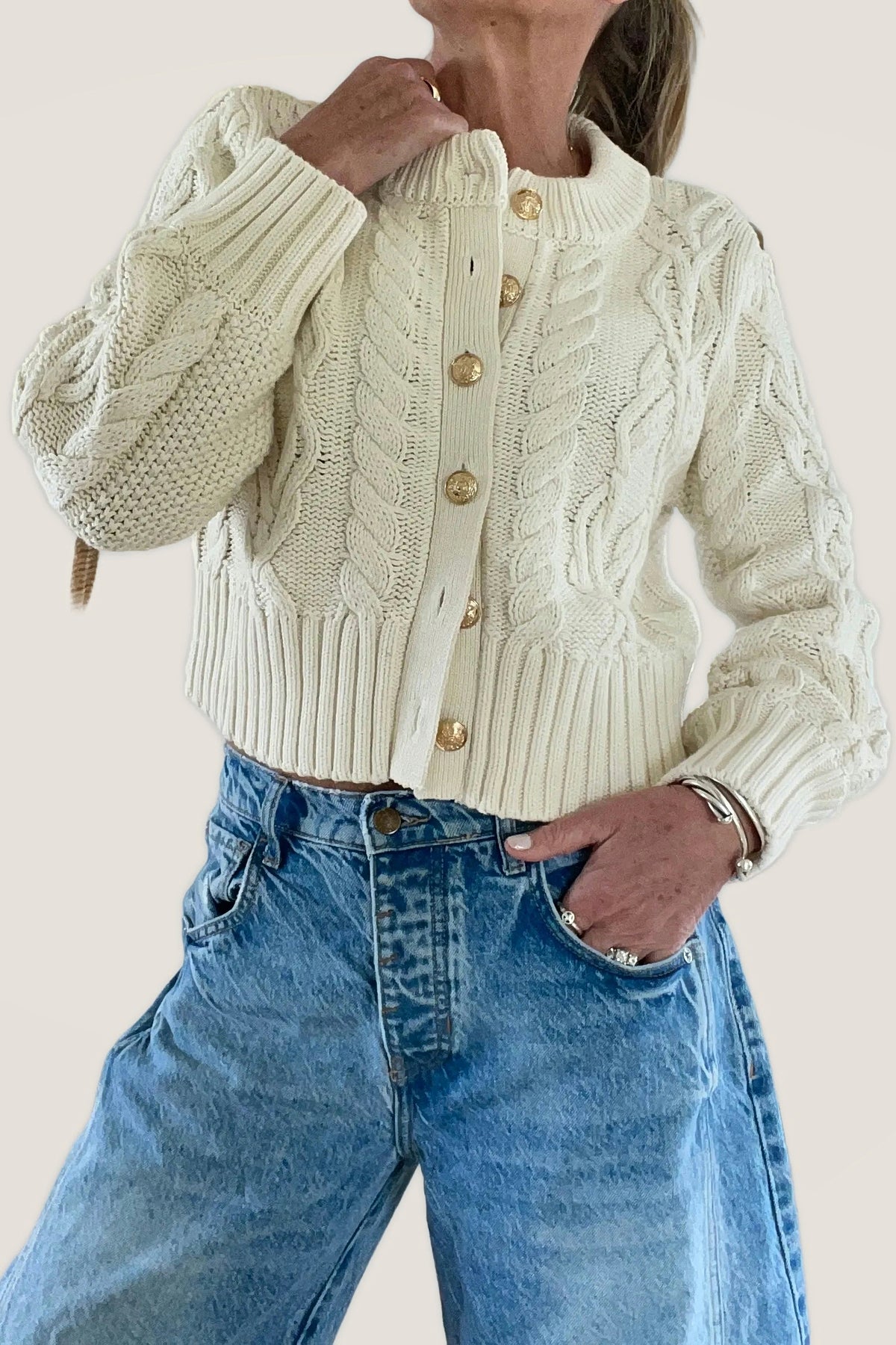 Front view of the ecru cable knit cardigan with gold crest buttons, worn with light wash jeans.