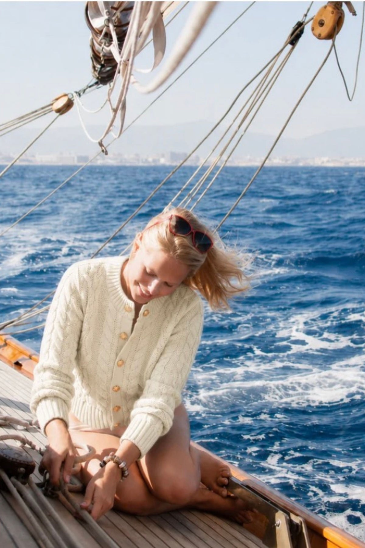 A woman sits on the wooden deck of a sailing boat, wearing a cream cable-knit cardigan and sunglasses on her head, with the blue ocean and distant coastline in the background.