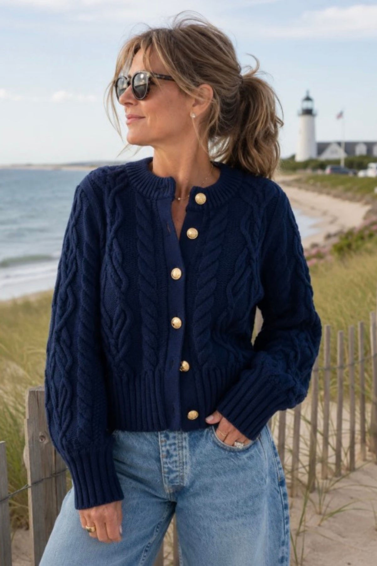 A woman wearing a navy blue cable-knit cardigan with gold buttons, paired with light blue jeans, standing on a dune path with a beach and lighthouse in the background.