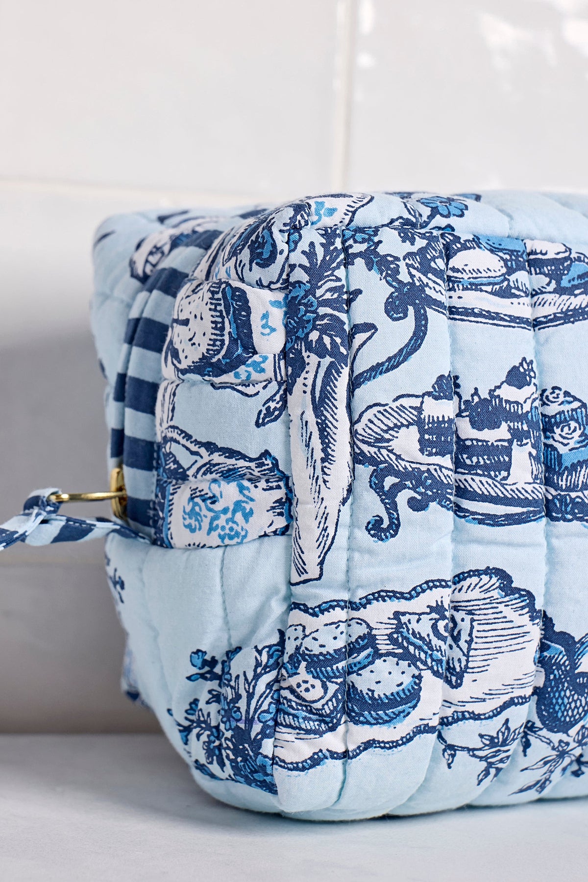 A close-up of the quilted High Tea toile pouch showing the blue print, brass zip and striped pull tab.
