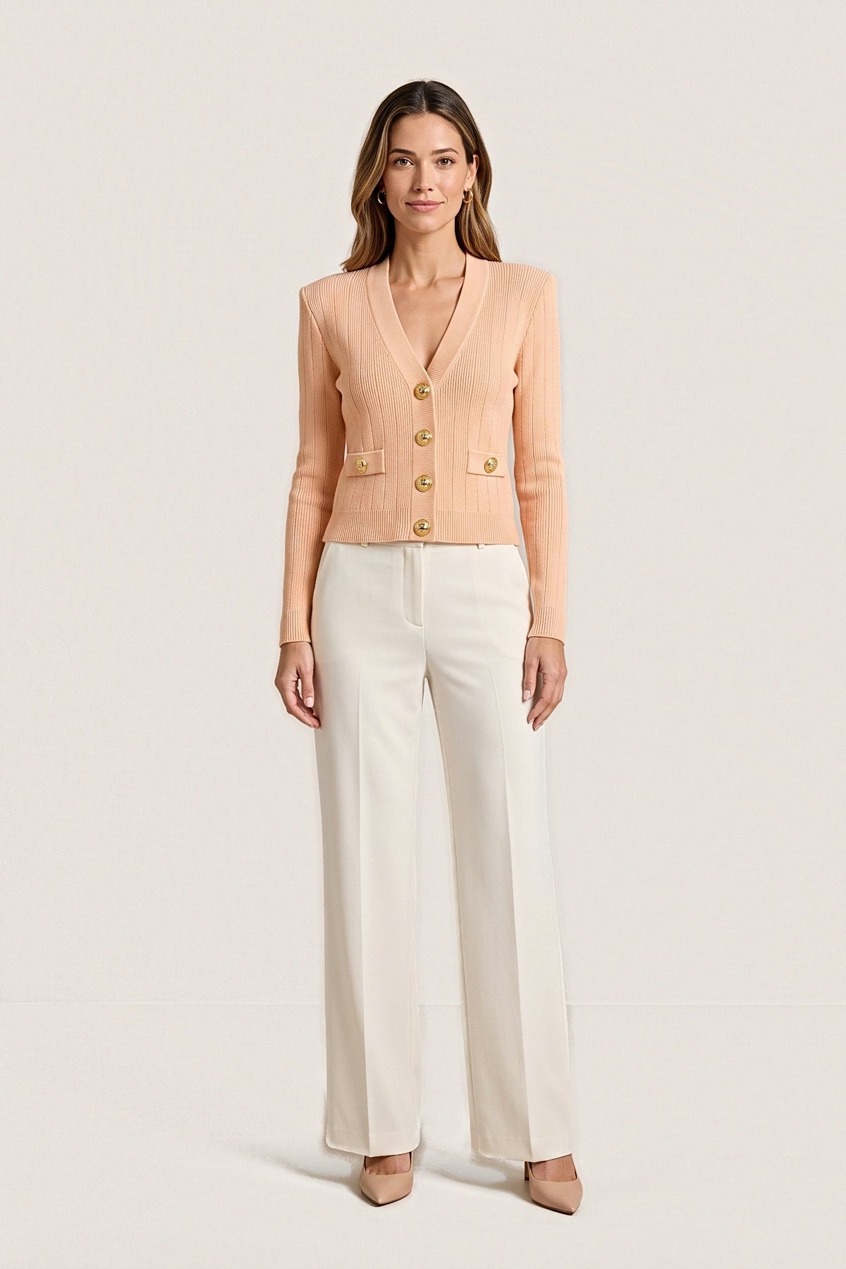 Front view of the Timeless Knit Cardigan in peach: a cropped ribbed V-neck cardigan with four gold buttons, two button-tab pockets and structured shoulders, worn with ivory wide-leg trousers.