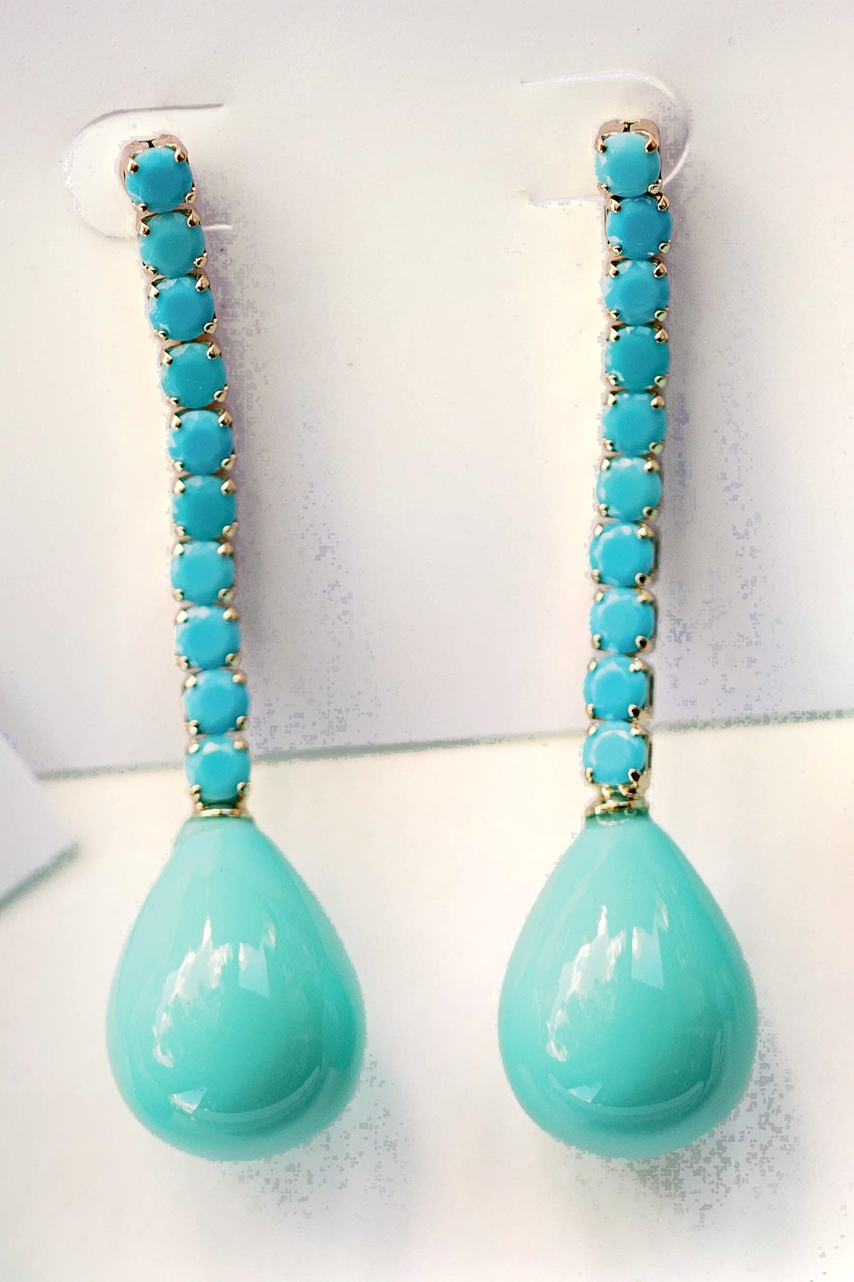 The turquoise Swingy statement earrings hanging from a display card, showing the line of turquoise stones set in gold and the teardrop.