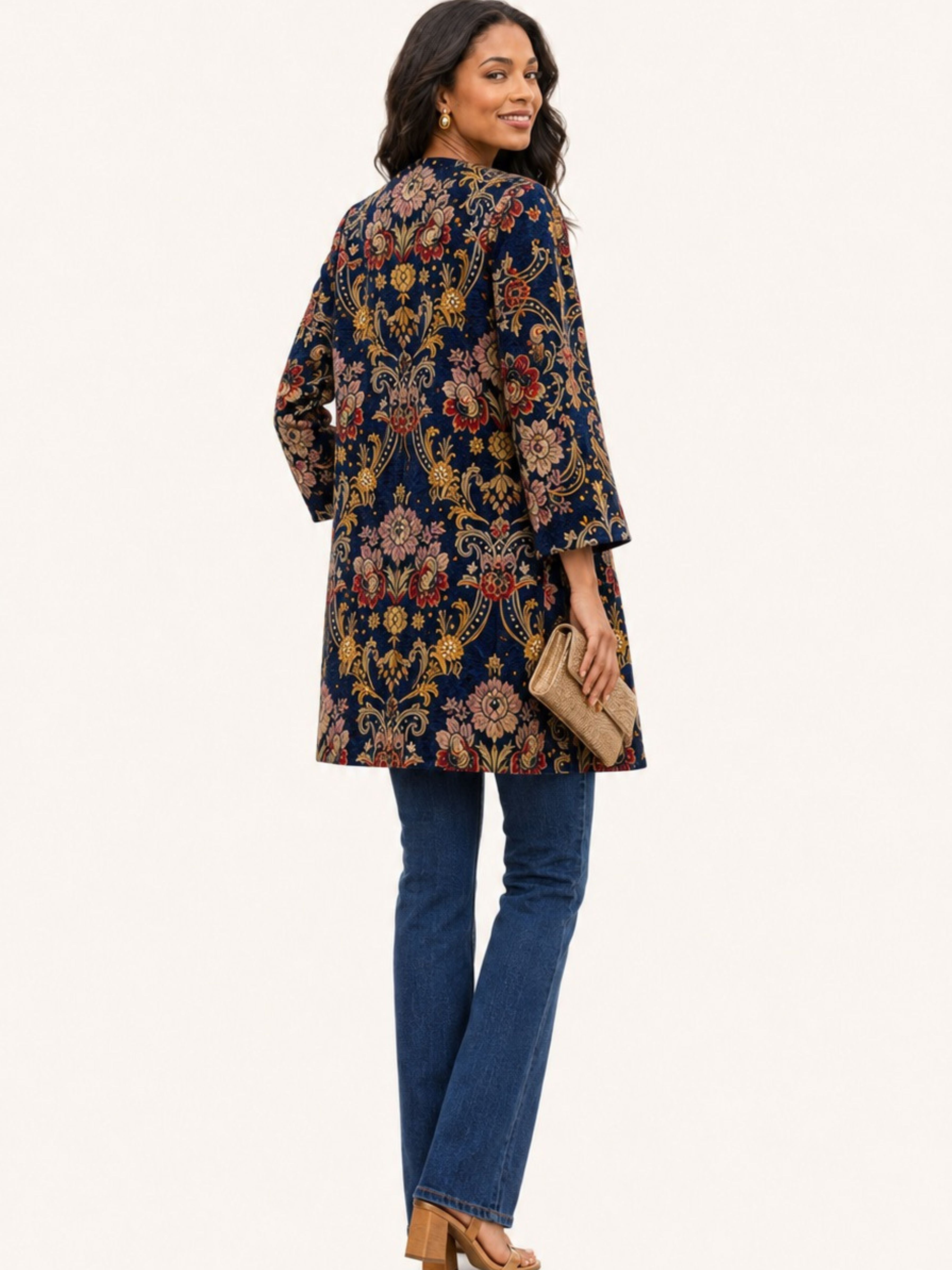 A woman modeling a navy blue tunic top with an intricate multicolor floral and paisley pattern, paired with blue jeans and holding a beige clutch bag against a plain off-white background.