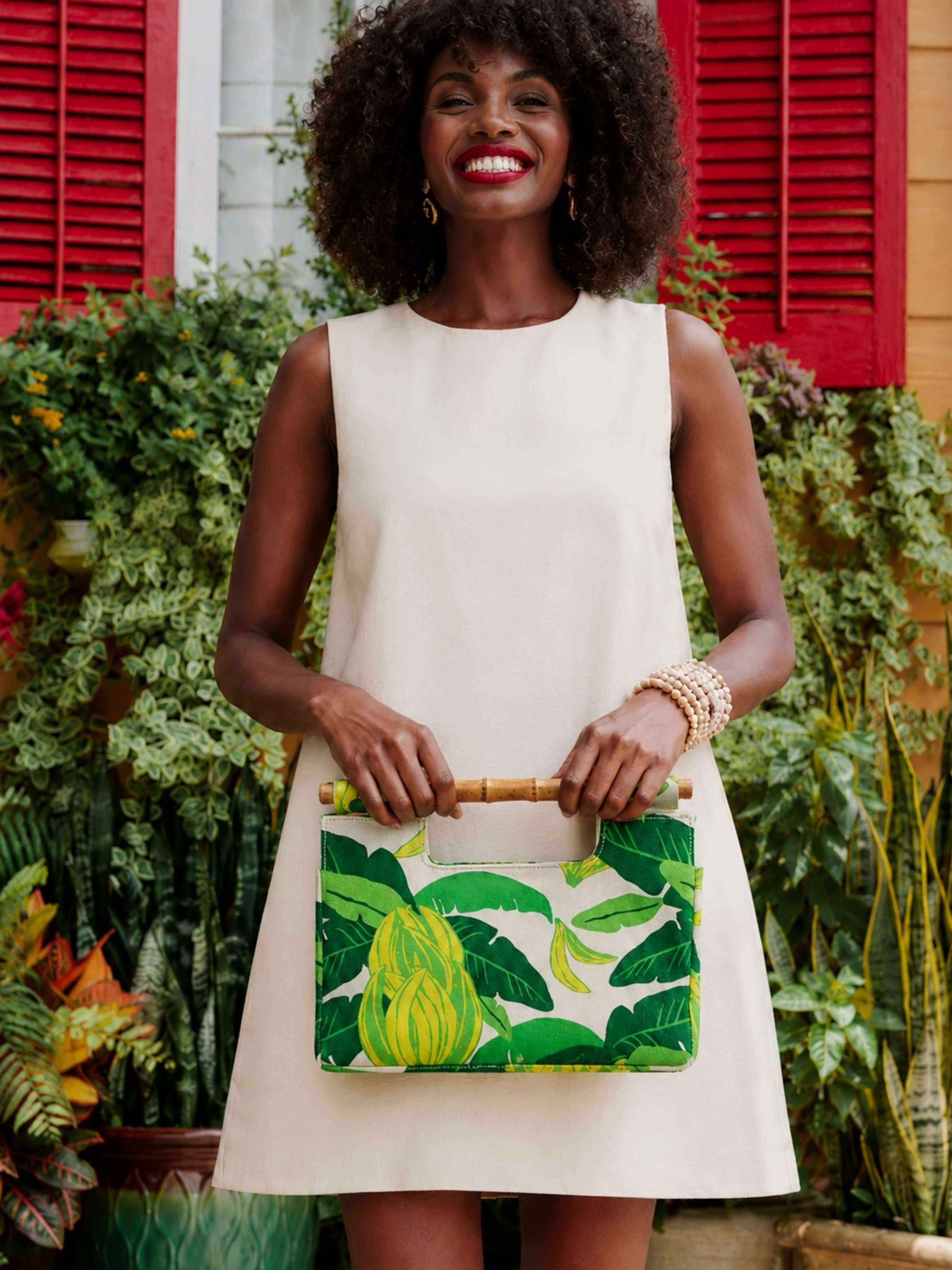 A woman with an afro hairstyle wearing a cream sleeveless dress and holding a green tropical leaf-patterned clutch bag with a bamboo handle, standing in front of a background of red shutters and green plants.