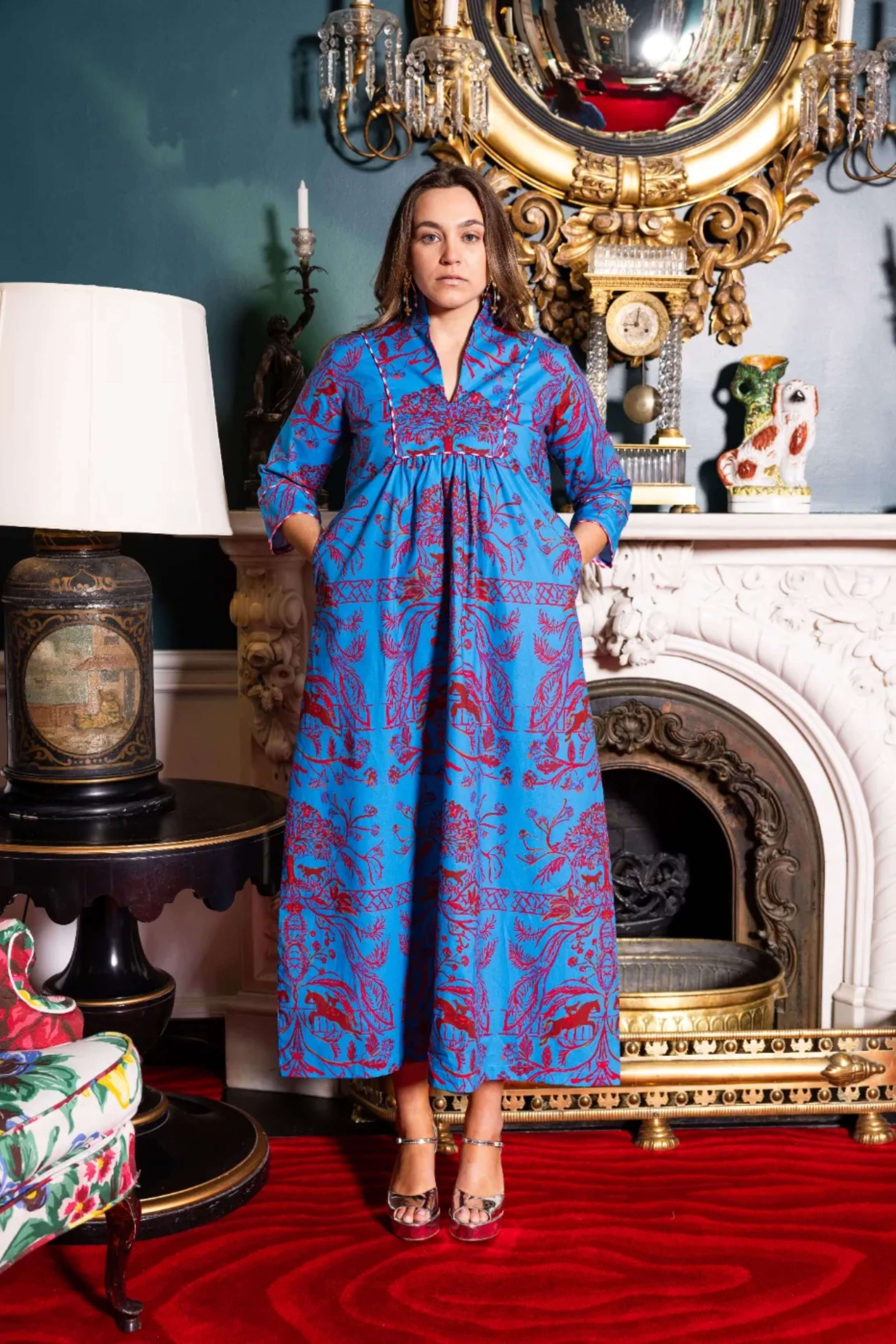Woman in a blue patterned dress standing in an ornate room with a fireplace and decorative mirror.