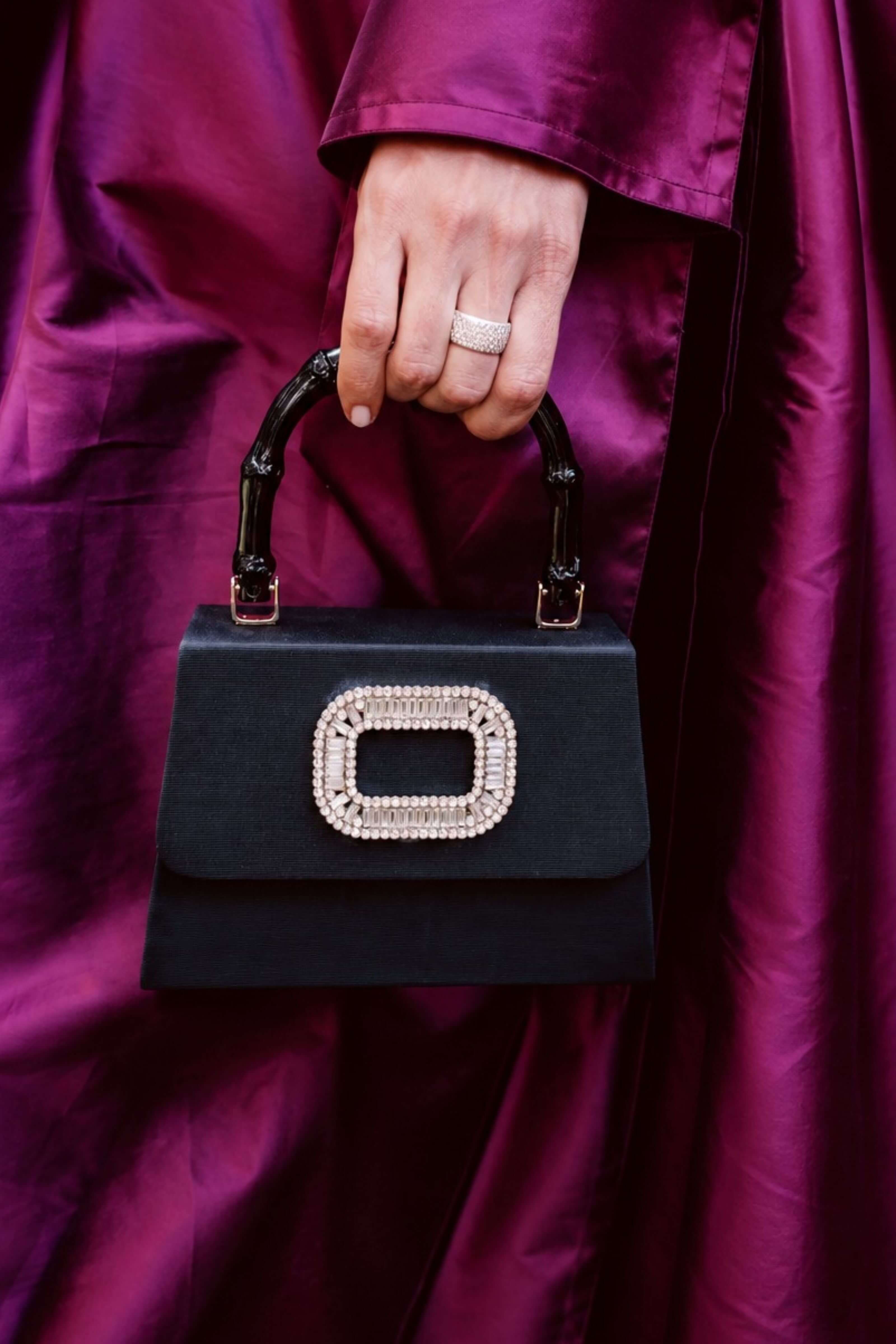 A close-up of a hand holding a structured black handbag with a large rhinestone-embellished rectangular clasp and a black bamboo-style top handle, set against a rich magenta satin garment.