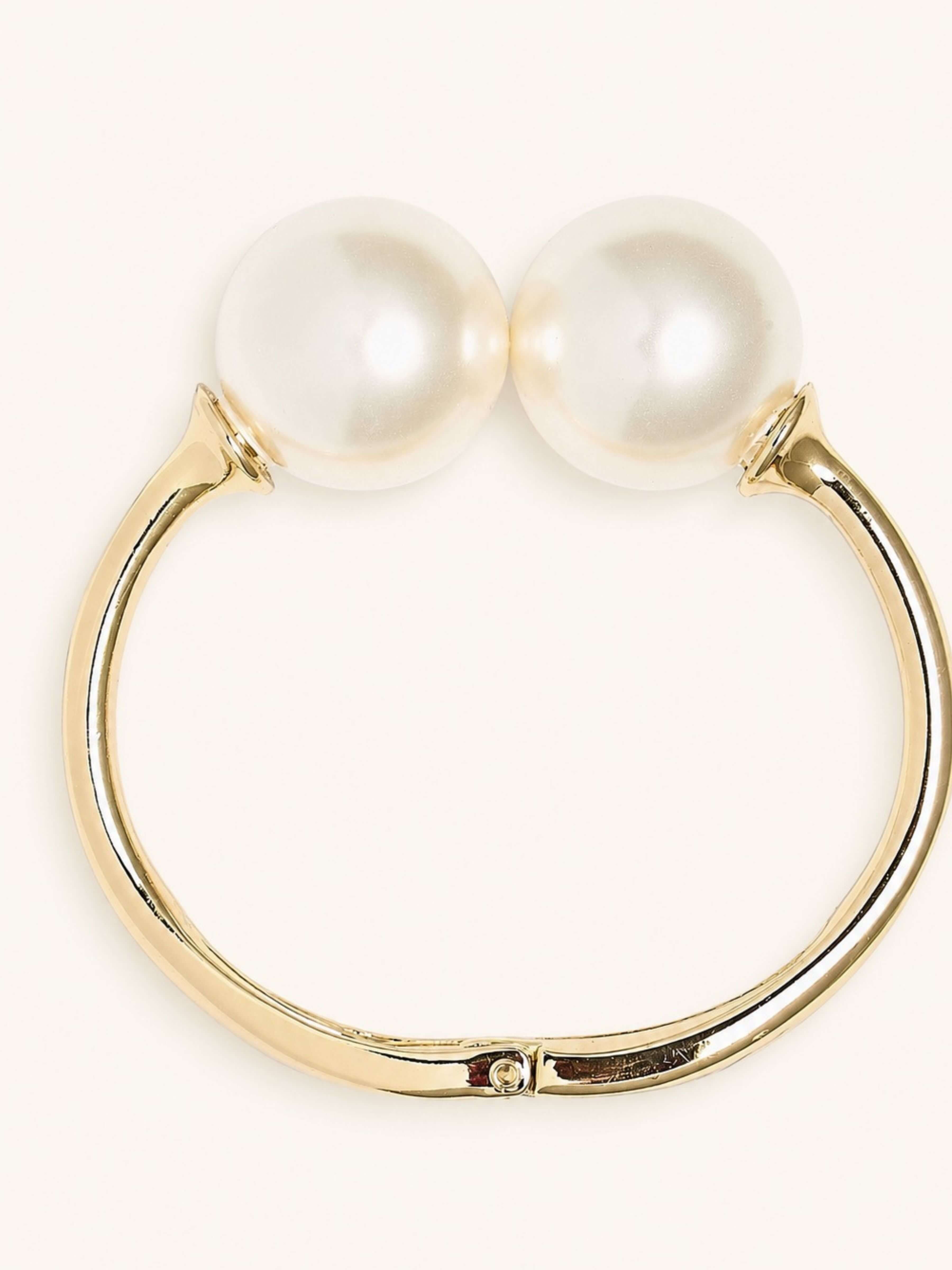 A gold-plated huggie ring featuring two large, round white faux pearls at the top, shown against a plain off-white background.
