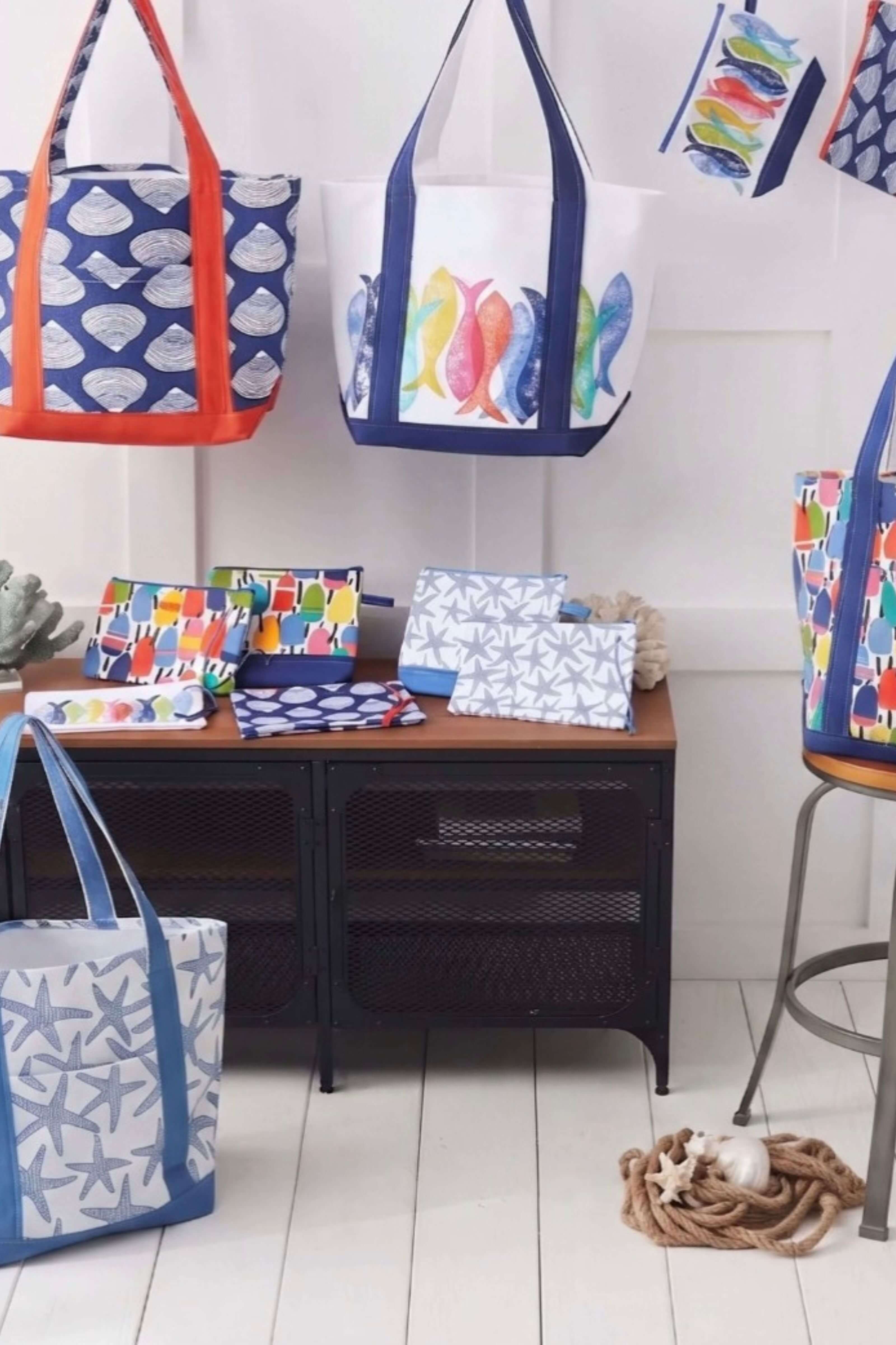 A collection of colorful beach tote bags and matching pouches displayed on a black metal side table in a bright, white-walled room. The bags feature various patterns including blue and white starfish, multicolor fish, and red and blue handles.