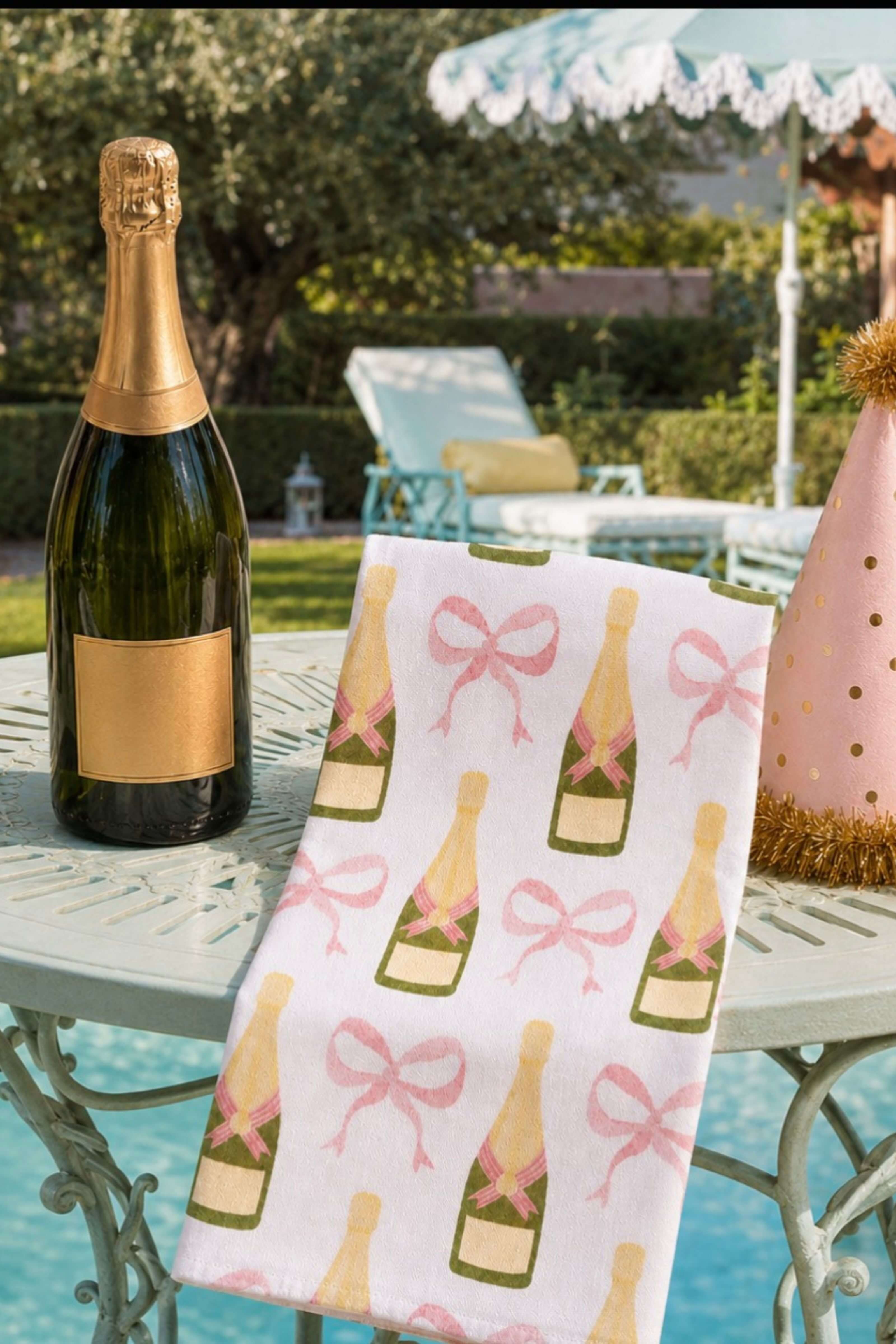 A white kitchen towel with a repeating champagne bottle and pink bow pattern, draped over a light green outdoor table next to a bottle of champagne in a poolside backyard setting.