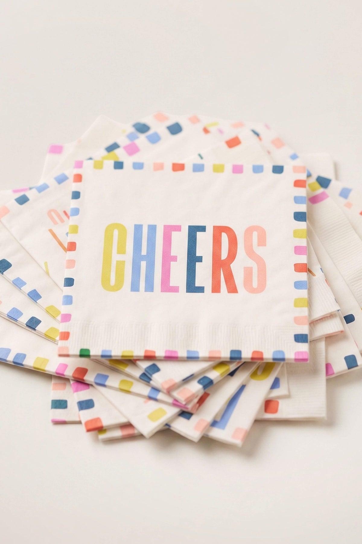 A stack of white paper party napkins with colorful confetti borders and the word 'CHEERS' printed in multicolored letters on the top napkin, photographed on a light neutral background.