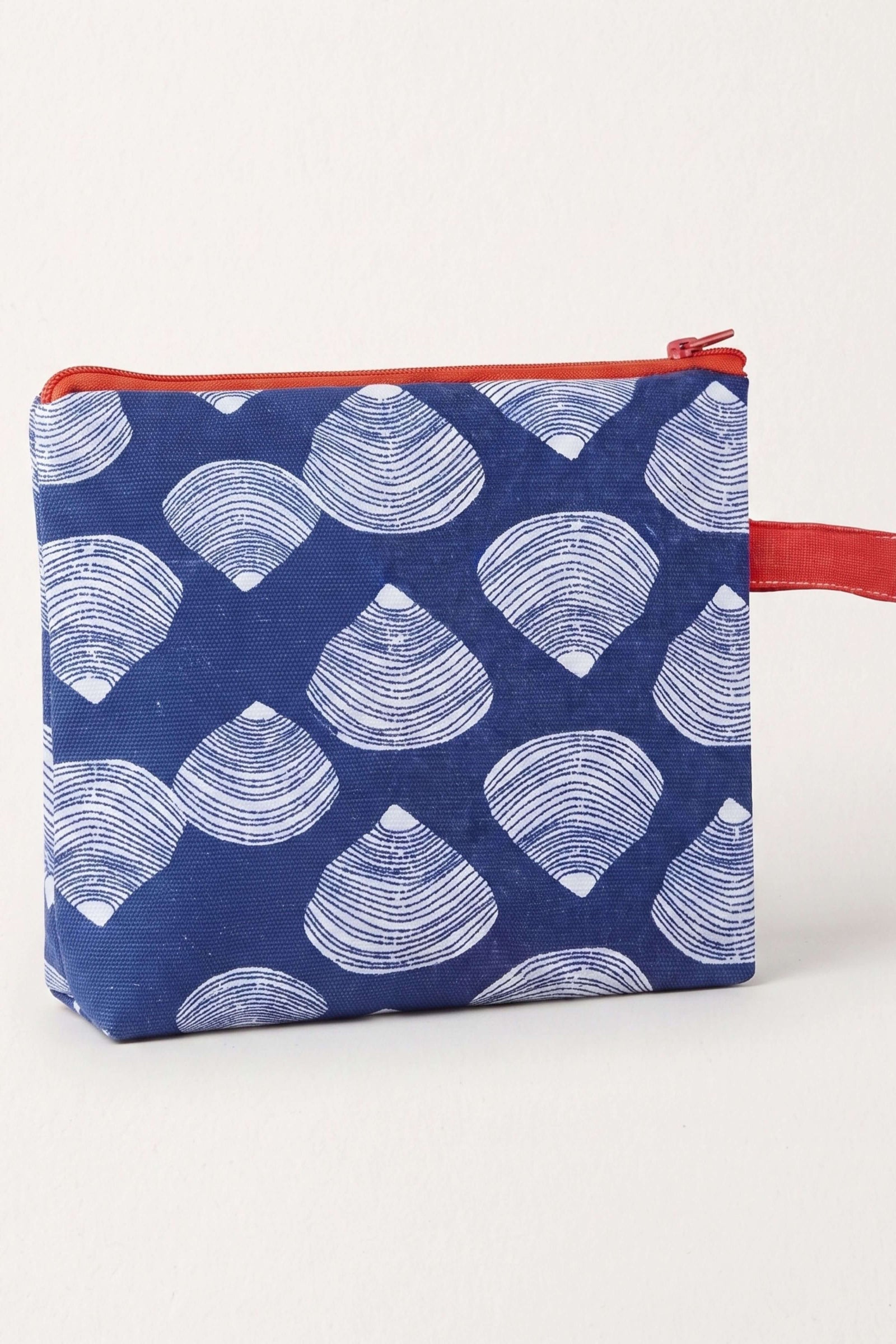 A small zippered pouch with a navy blue background and a repeating white scallop shell pattern, featuring a red zipper and a red wrist strap, shown on a light off-white background.