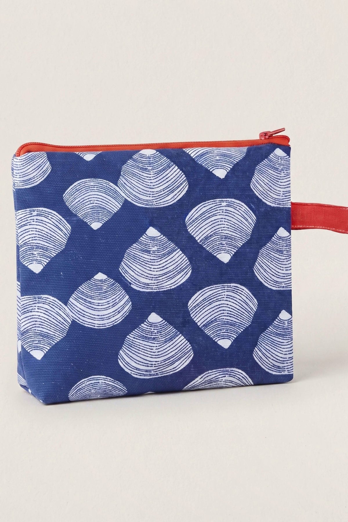 A small zippered pouch with a navy blue background and a repeating white scallop shell pattern, featuring a red zipper and a red wrist strap, shown on a light off-white background.