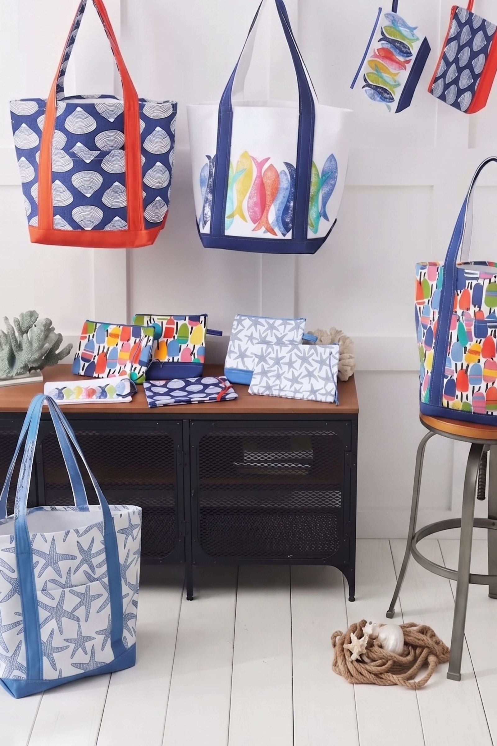 A collection of colorful beach tote bags and matching zip pouches displayed on a white wall and floor, featuring various nautical prints like starfish, fish, and seashells.