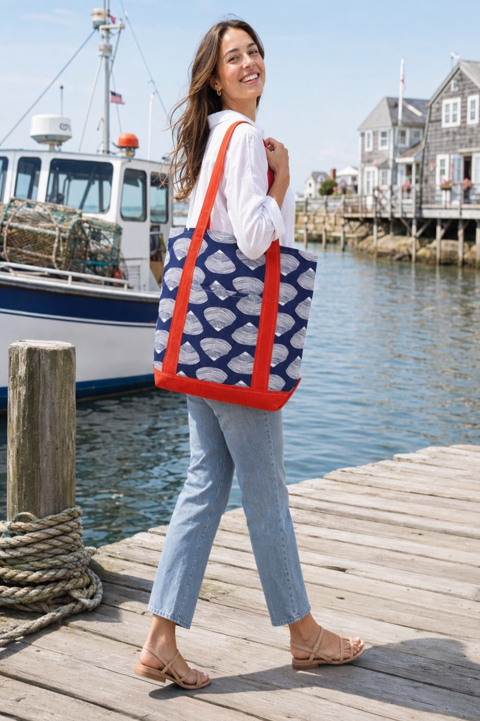 A woman walking on a wooden dock by the water wearing a navy blue tote bag with a white fish pattern and bright red straps. A fishing boat and coastal houses are visible in the background.
