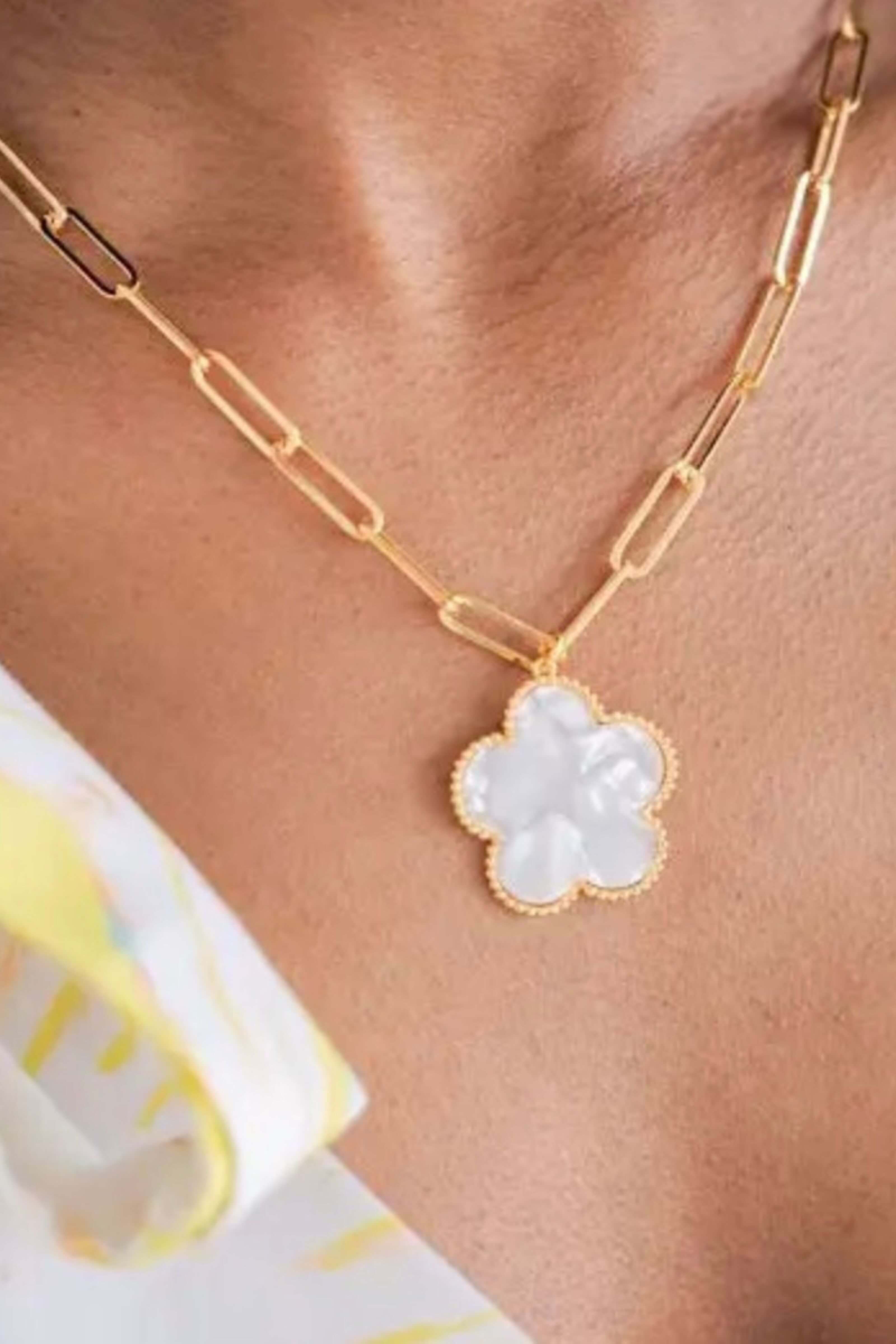 A close-up of a gold paperclip chain necklace with a white clover-shaped pendant, worn on a model's neck.