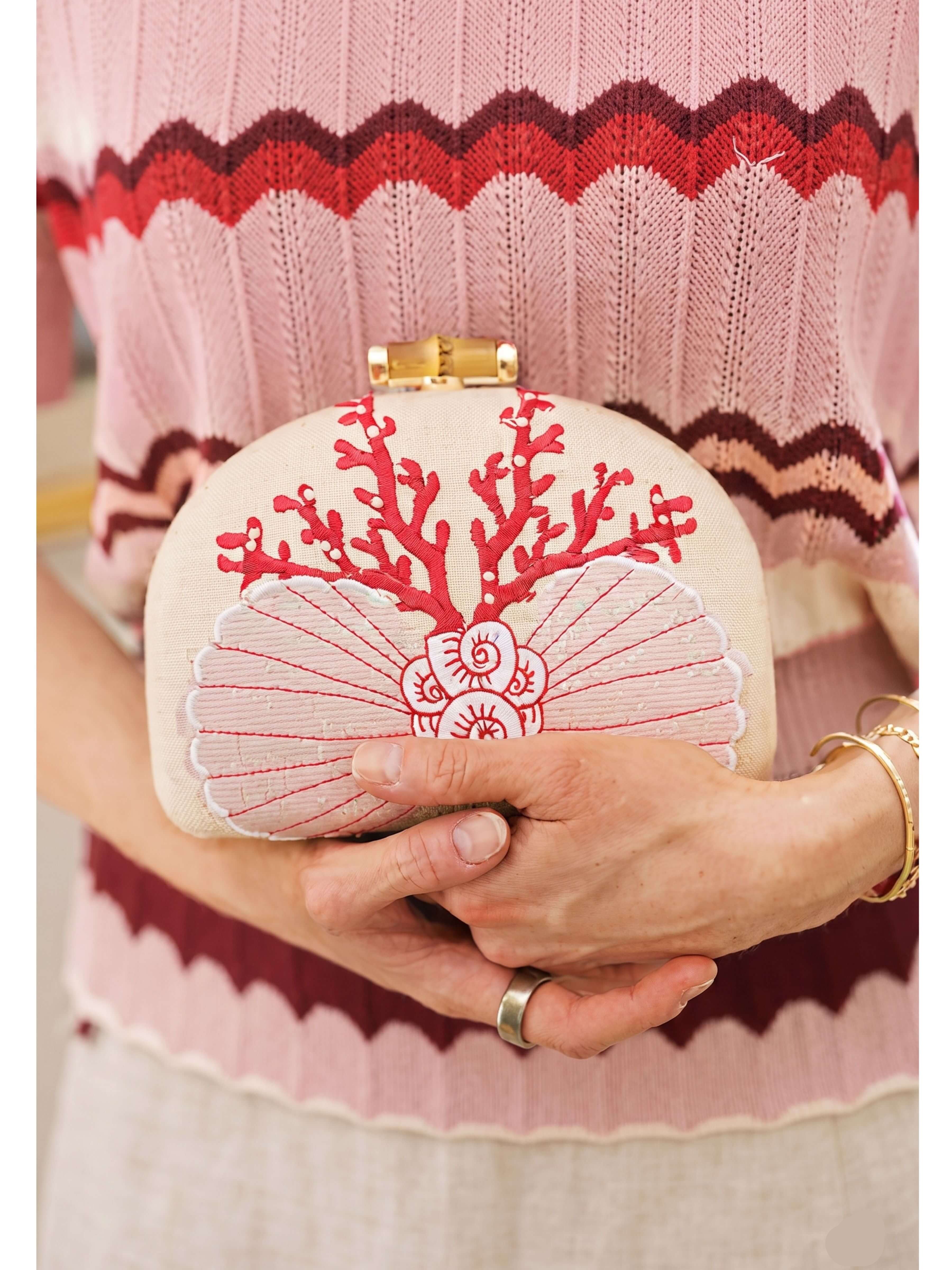A person in a pink and burgundy chevron-knit sweater holds a small oval clutch bag. The clutch is cream-colored with a red coral-like embroidery on top and a pink seashell embroidery on the front, finished with a gold-tone clasp.