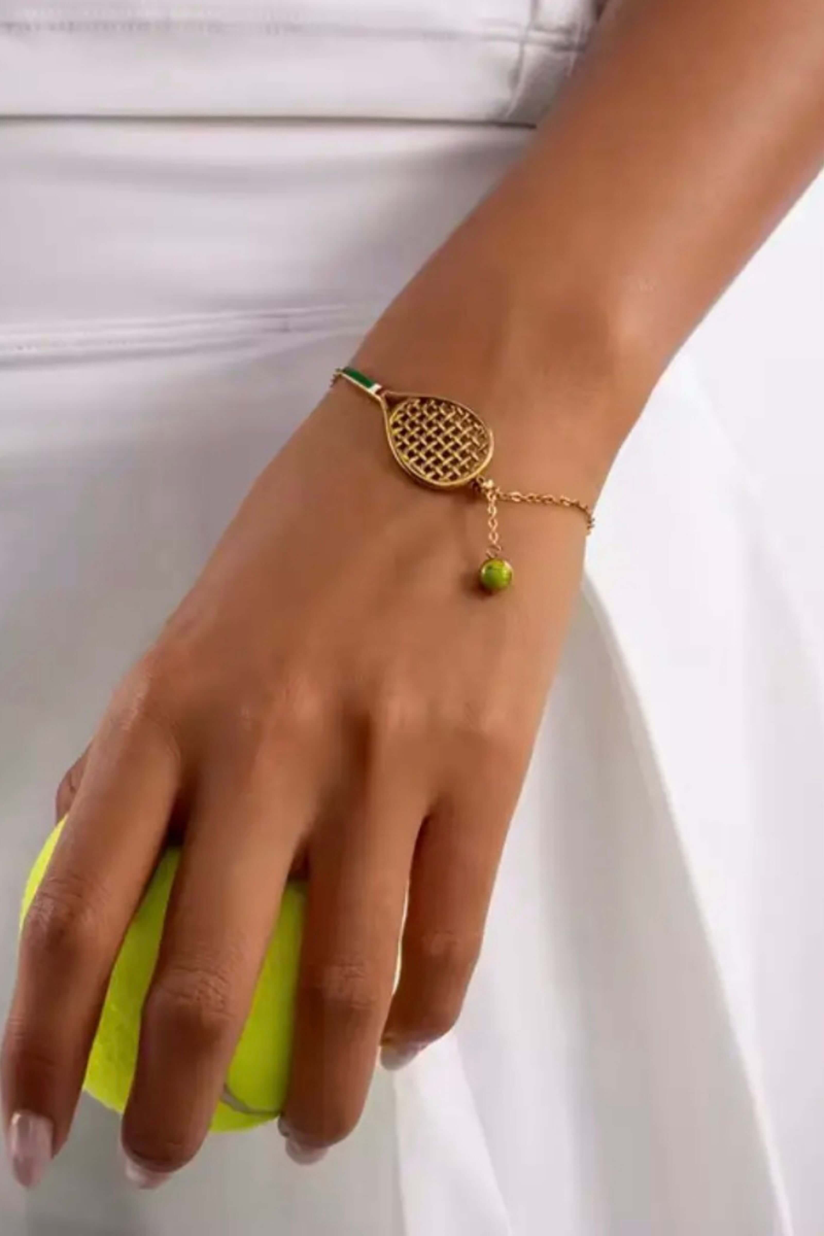 A gold tennis bracelet with a tennis racket charm and a small green bead, worn on a woman's wrist while she holds a yellow tennis ball against a white tennis dress background.