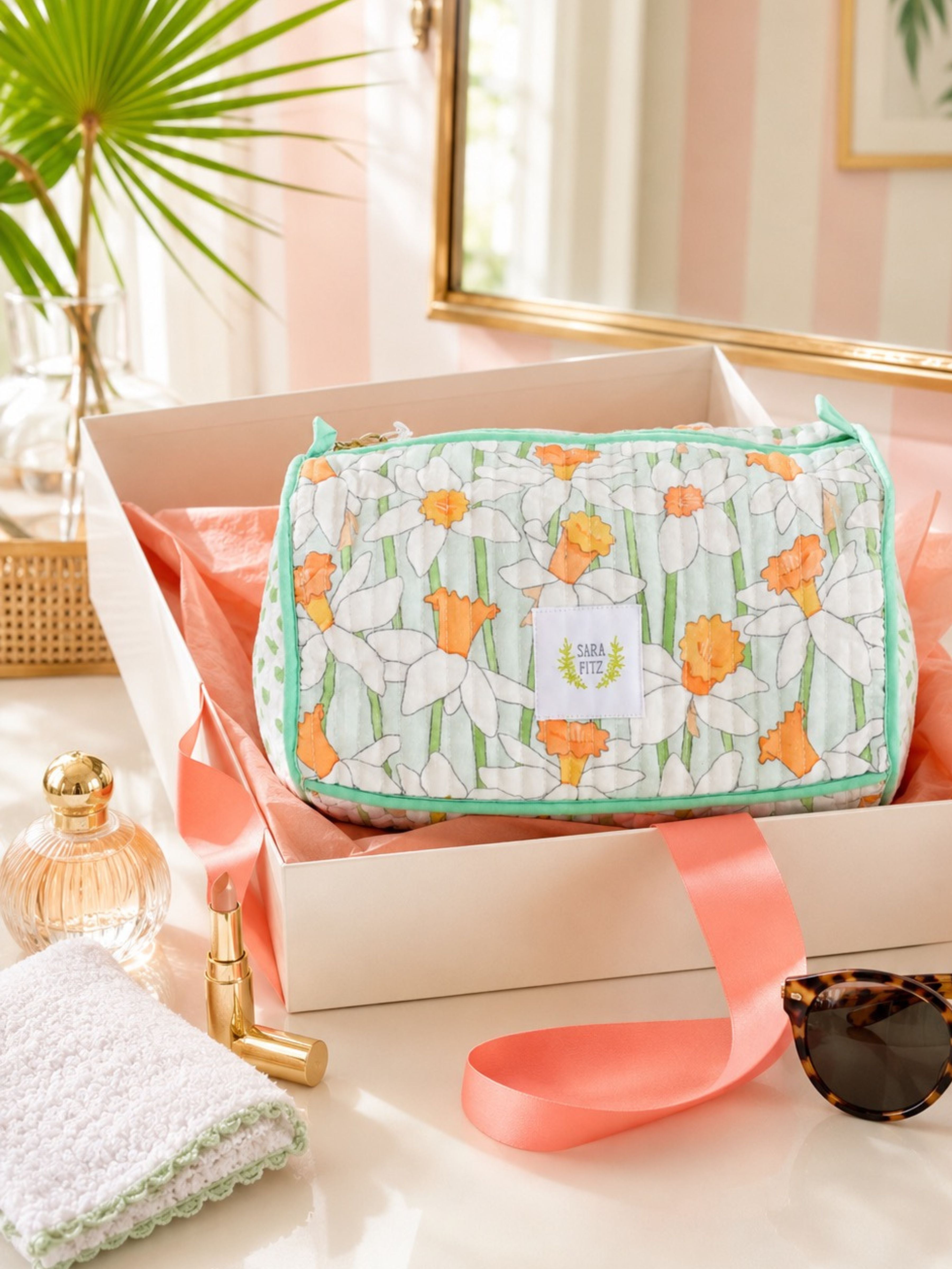 A cosmetic pouch with a daffodil floral print in white, green, and orange, presented inside a white gift box with pink ribbon. The pouch features a white label with the brand name 'Sunny Day'.